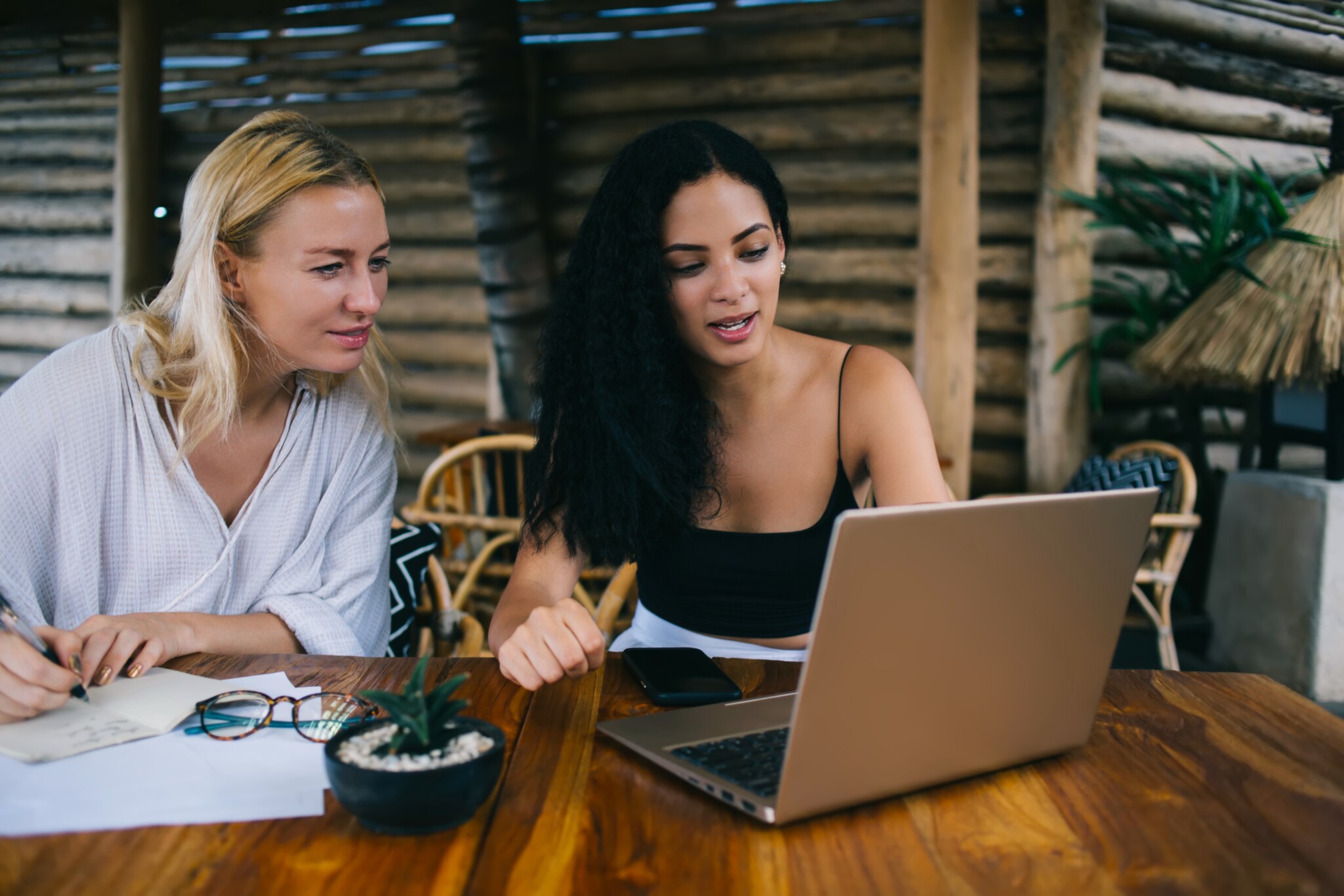 Zwei Frauen in Sommerkleidung sitzen an einem Holztisch vor einem Laptop.