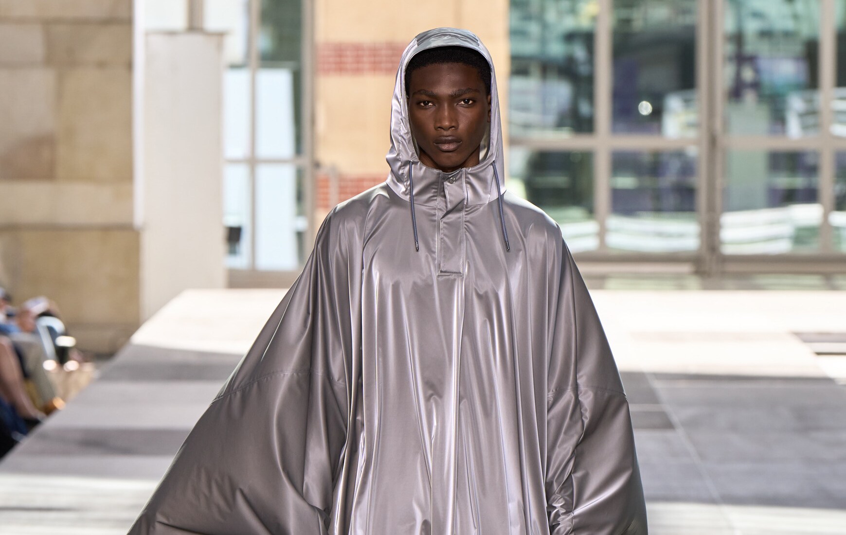 Ein Model im silberfarbenen Regencape läuft über einen Laufsteg direkt auf die Kamera zu