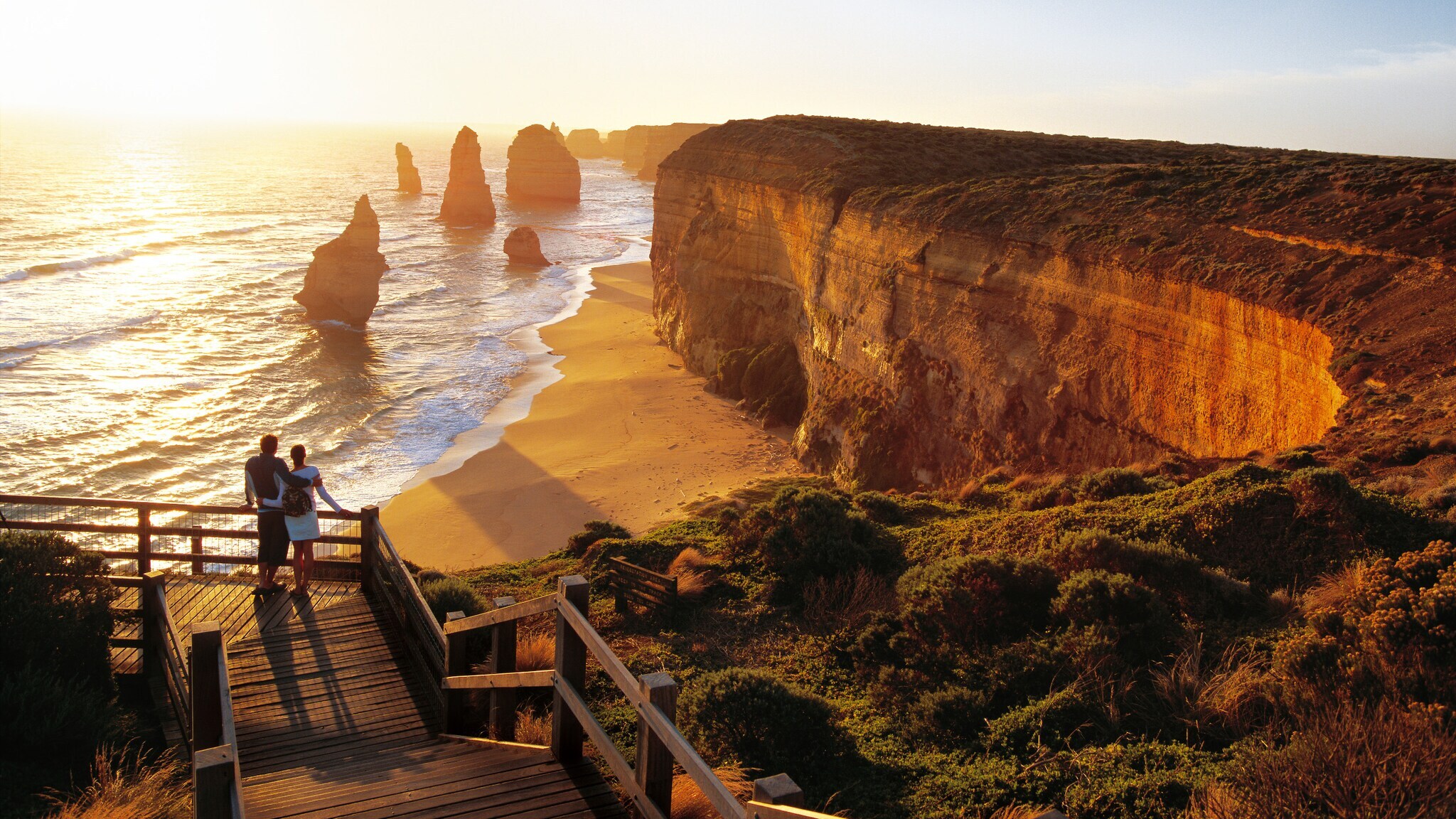 Ein Paar schaut von einer Aussichtsplattform auf eine Küste mit Felsformationen bei Sonnenuntergang