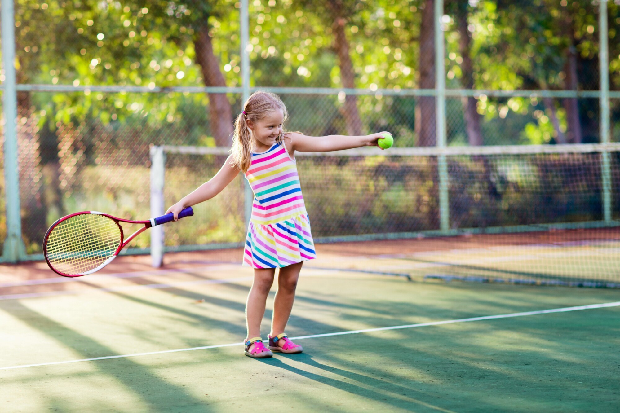 Ein kleines Kind im bunt gestreiften Kleid steht auf einem Tennisplatz und hält in der einen Hand einen Tennisschläger und in der anderen einen Tennisball.