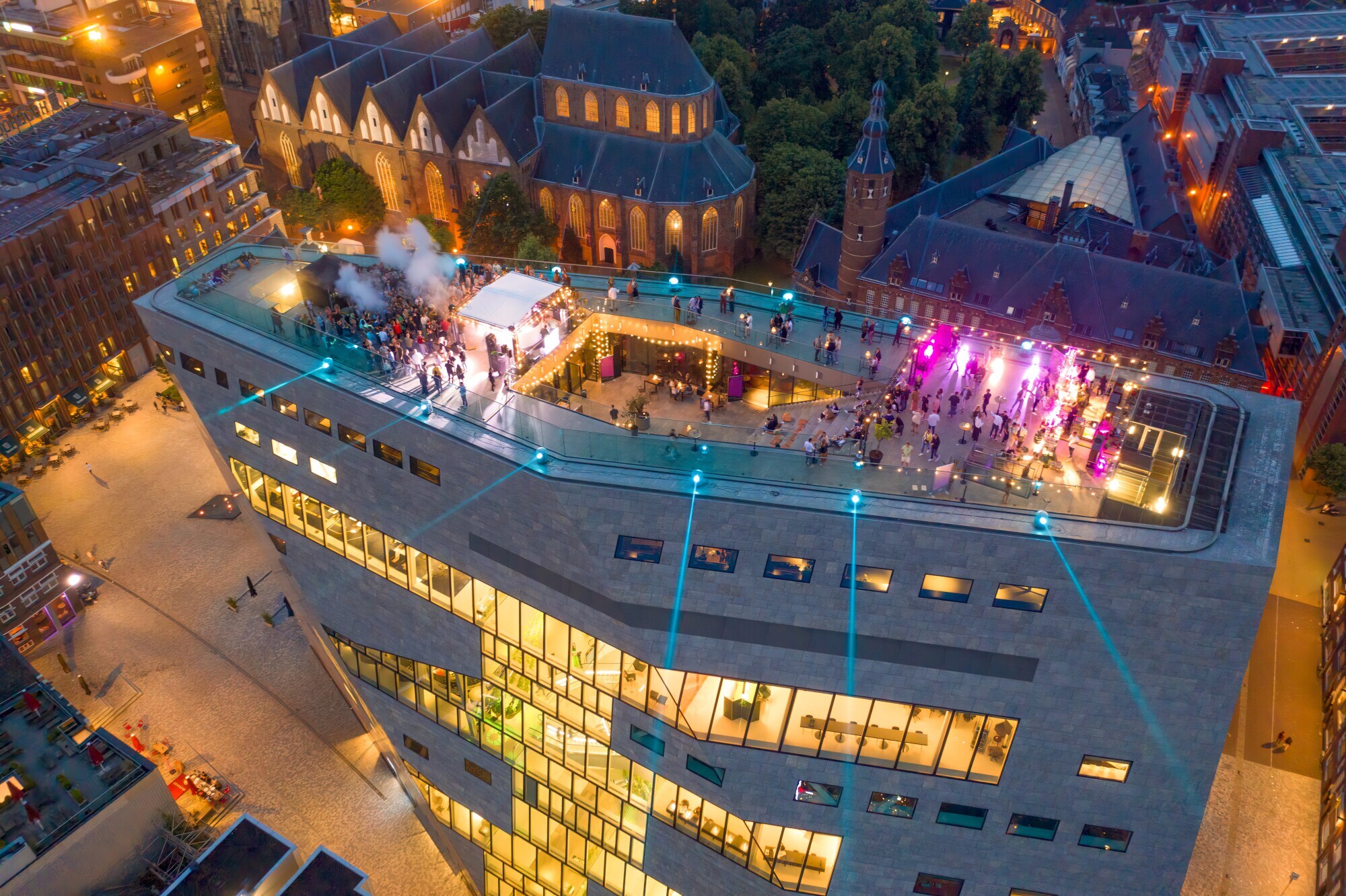 Luftaufnahme einer Veranstaltung auf einer großen Dachterrasse eines modernen, städtischen Gebäudekomplexes bei Nacht