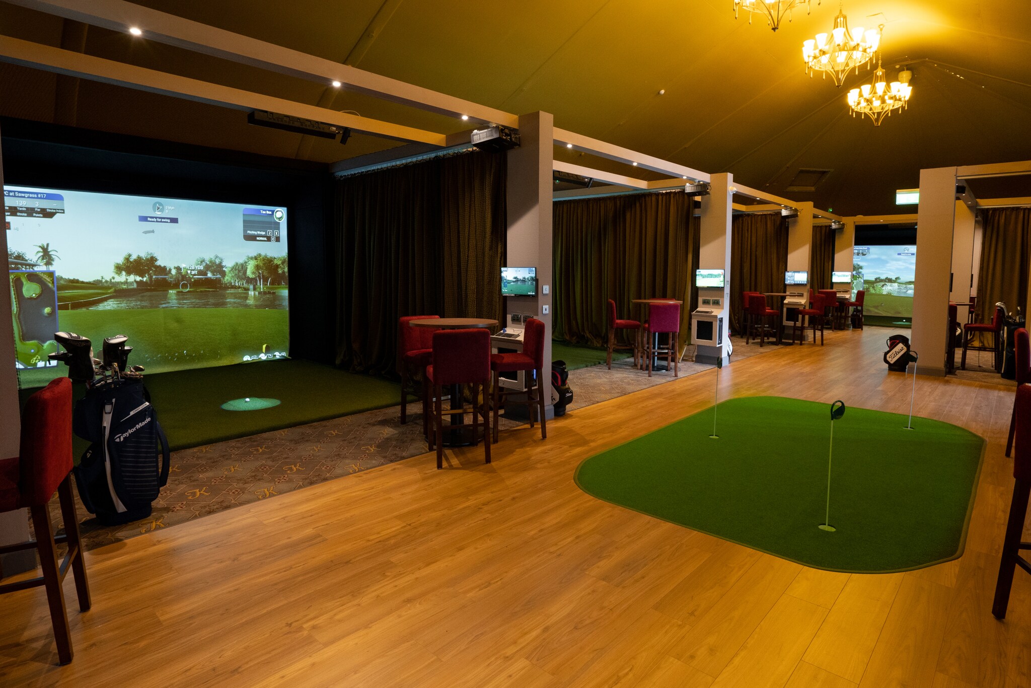 Ein großer Raum mit Holzboden, in dem mehrere Kabinen nebeneinander stehen, in denen ein digitaler Golfplatz gezeigt wird.