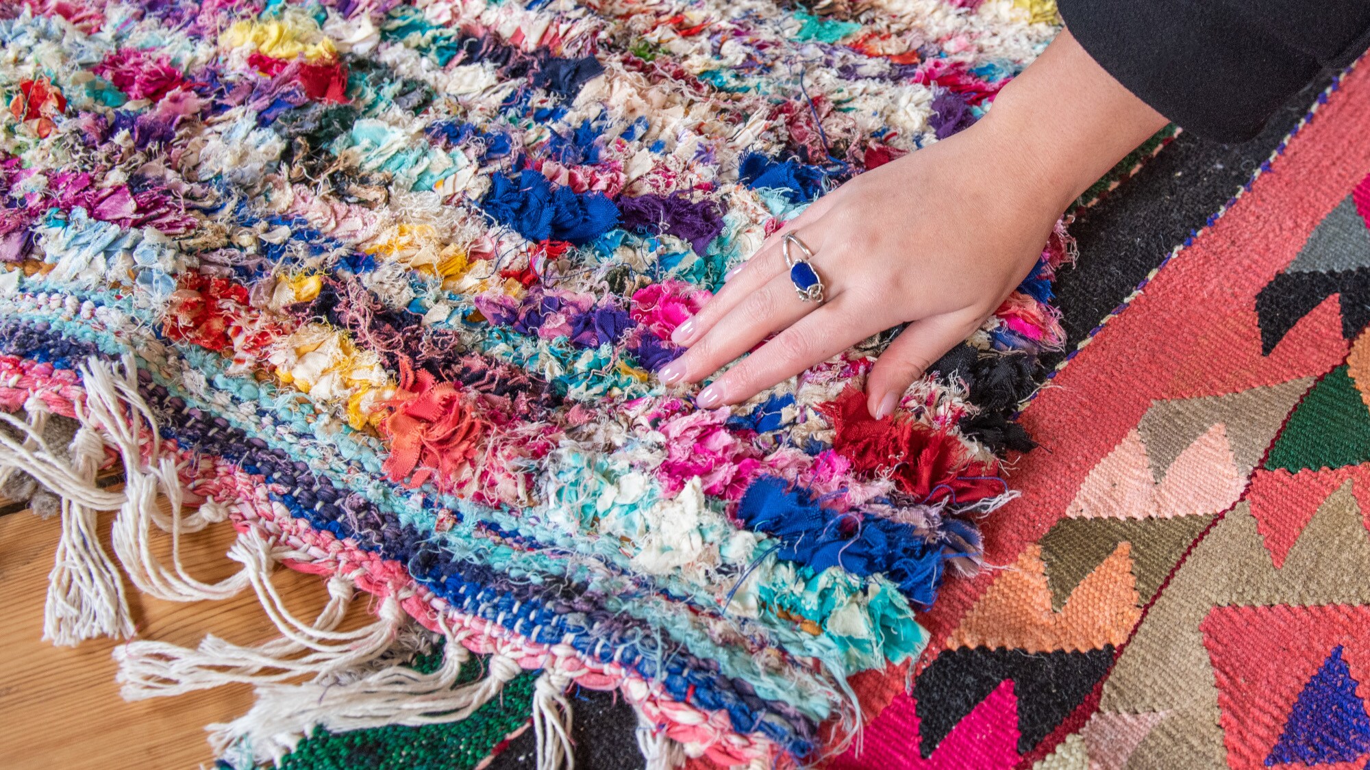 Detailaufnahme eines bunten Teppichs über den eine Hand mit blauem Ring streicht Detailaufnahme eines bunten Teppichs über den eine Hand mit blauem Ring streicht