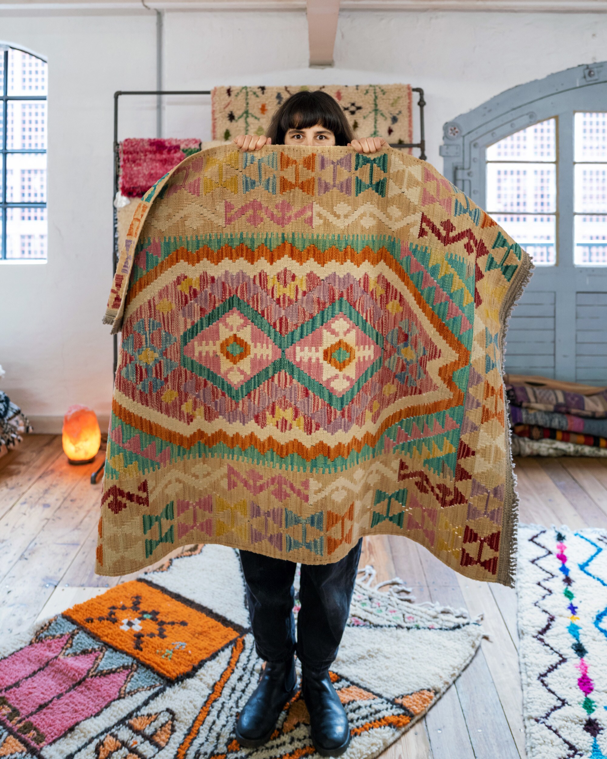 Anna Wahdat, die sich einen bunten Kelim vor das Gesicht hält, auf dem Boden liegen weitere Teppiche