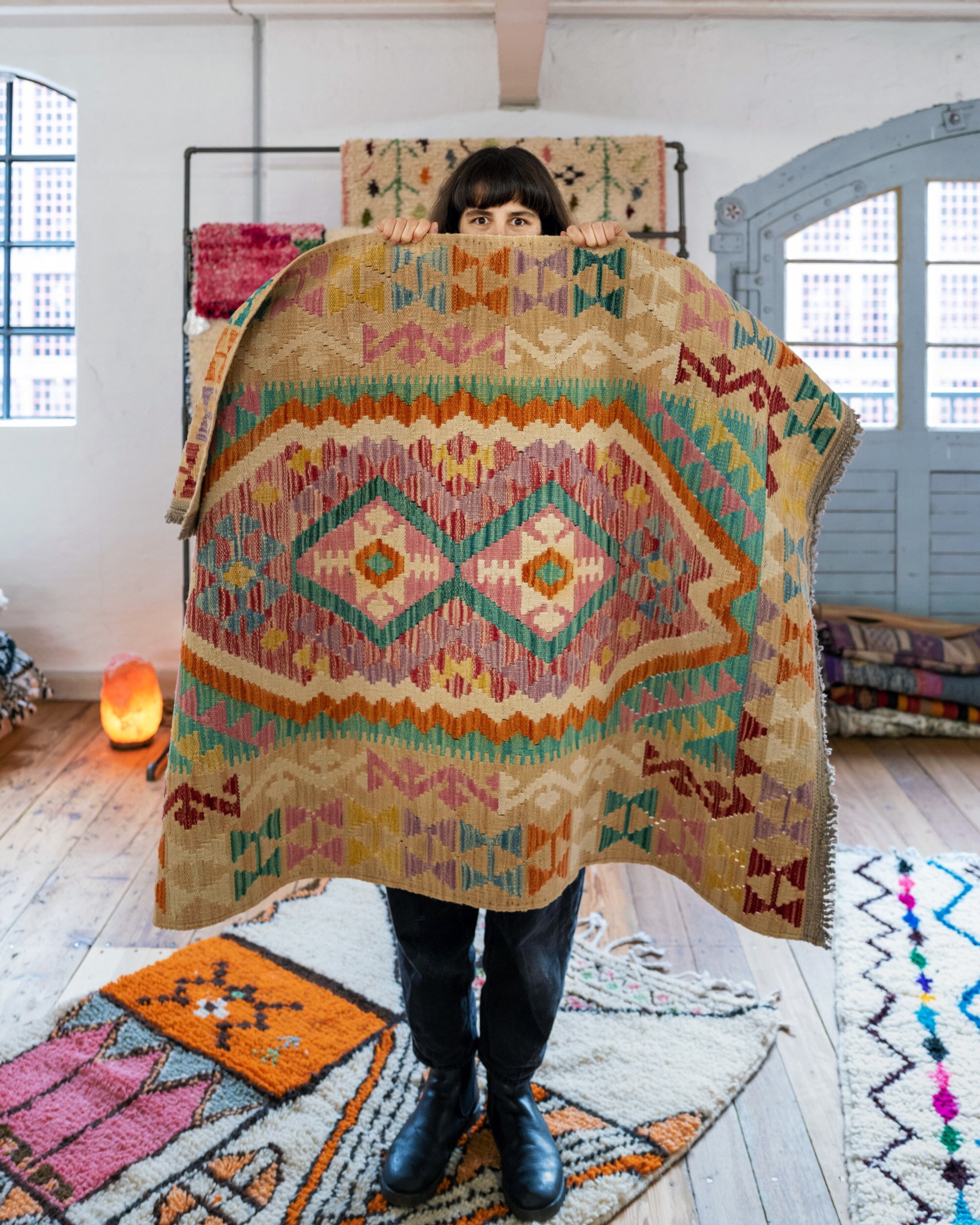 Anna Wahdat, die sich einen bunten Kelim vor das Gesicht hält, auf dem Boden liegen weitere Teppiche Anna Wahdat, die sich einen bunten Kelim vor das Gesicht hält, auf dem Boden liegen weitere Teppiche