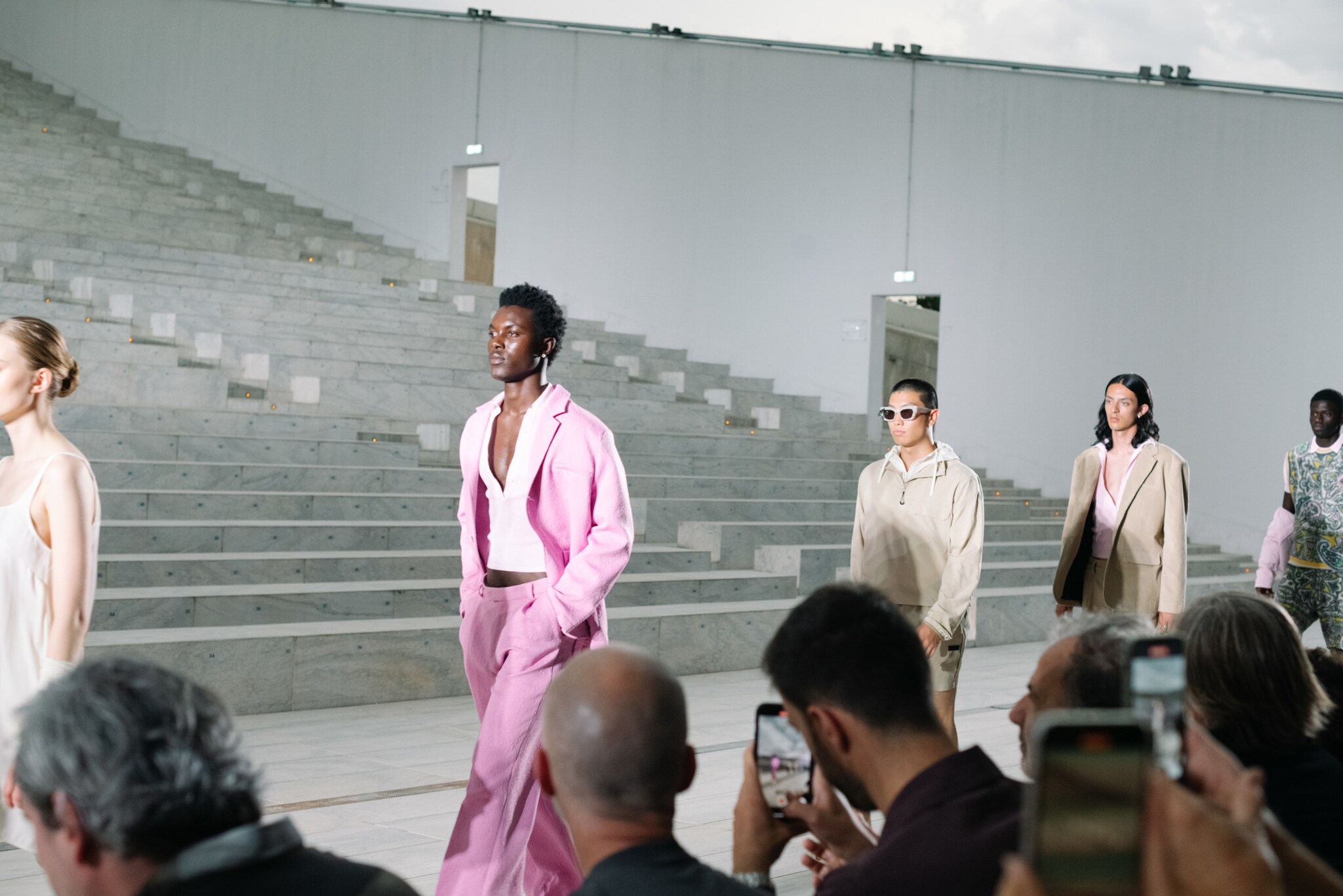 Männliche Models laufen bei einer Fashionshow, im Vordergrund sitzt das Publikum und filmt mit Handys die Show