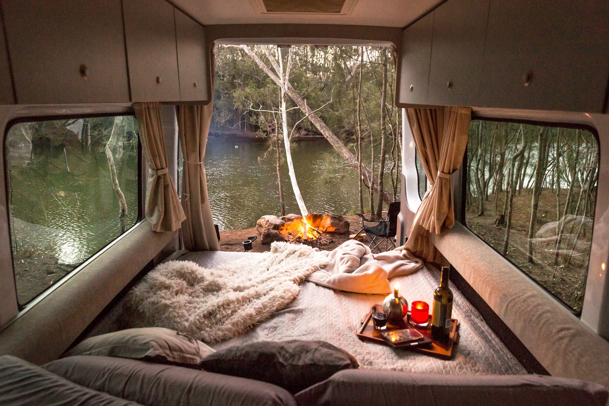 Innenansicht eines Campers an einem Fluss, davor ein Lagerfeuer Innenansicht eines Campers an einem Fluss, davor ein Lagerfeuer
