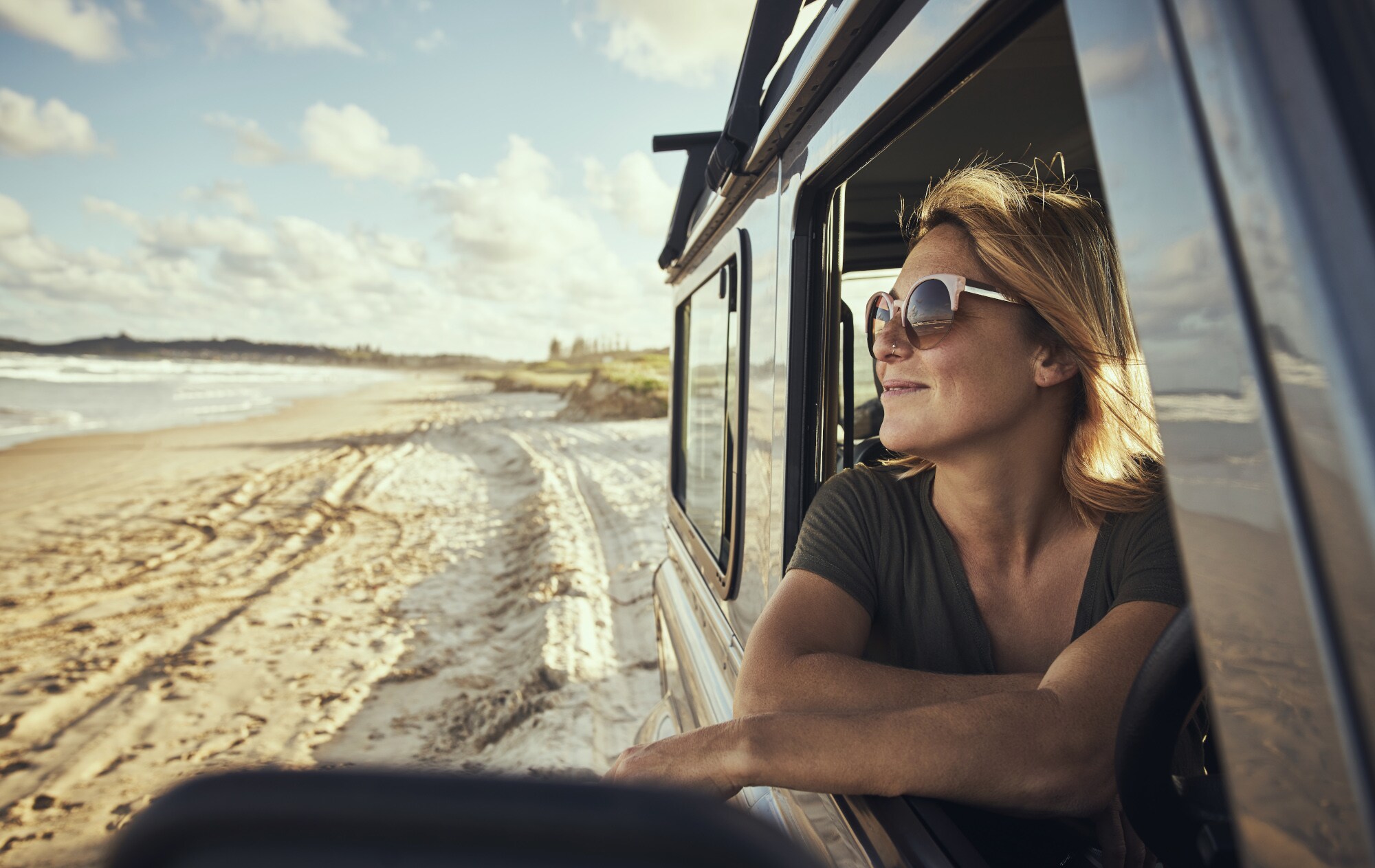 Eine lächelnde Frau mit Sonnenbrille in einem Auto am Strand Eine lächelnde Frau mit Sonnenbrille in einem Auto am Strand