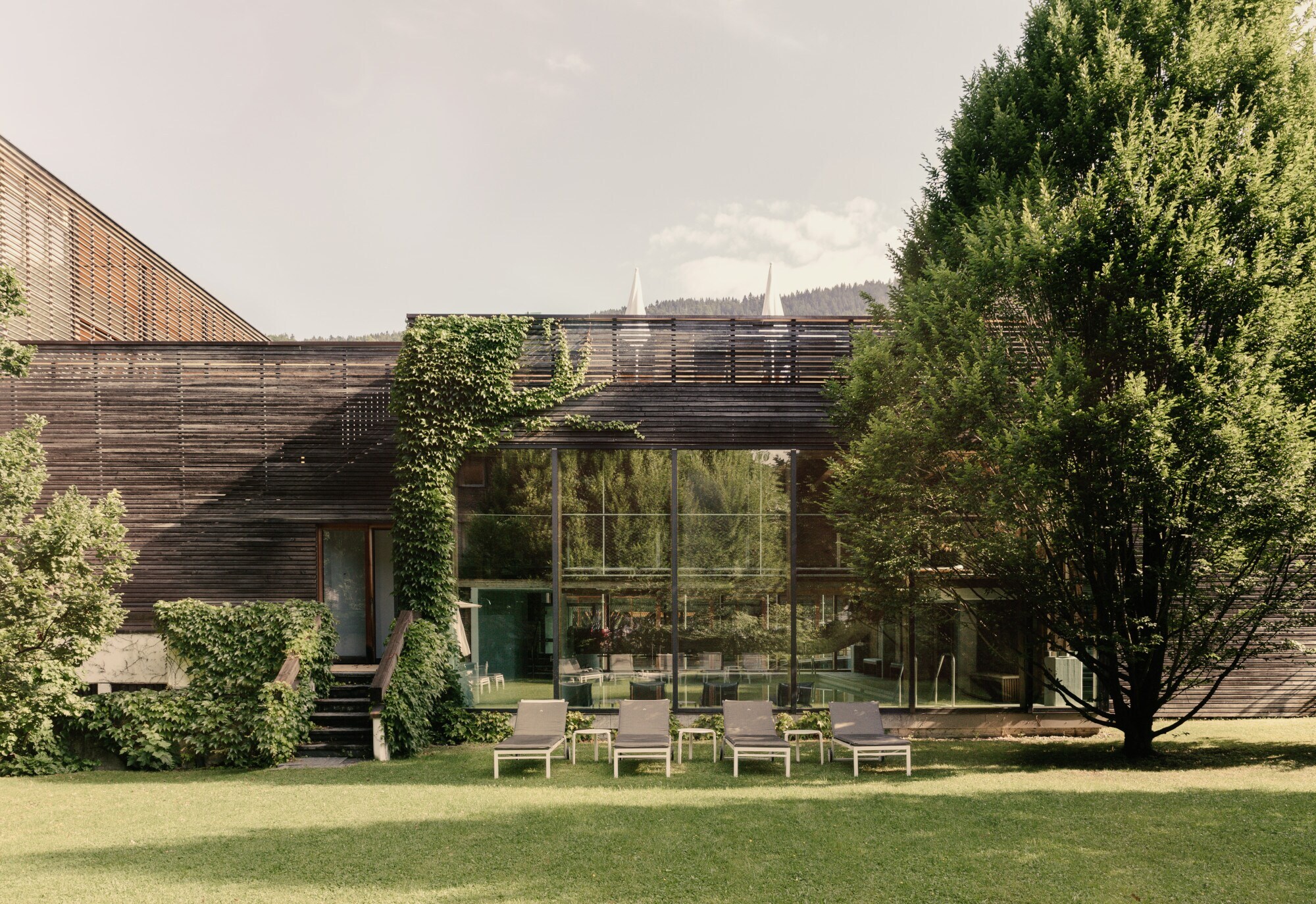 Blick auf ein modernes, holzverkleidetes Gebäude in dessen Inneren ein Swimmingpool zu sehen ist, im Vordergrund Liegestühle und Grünfläche.