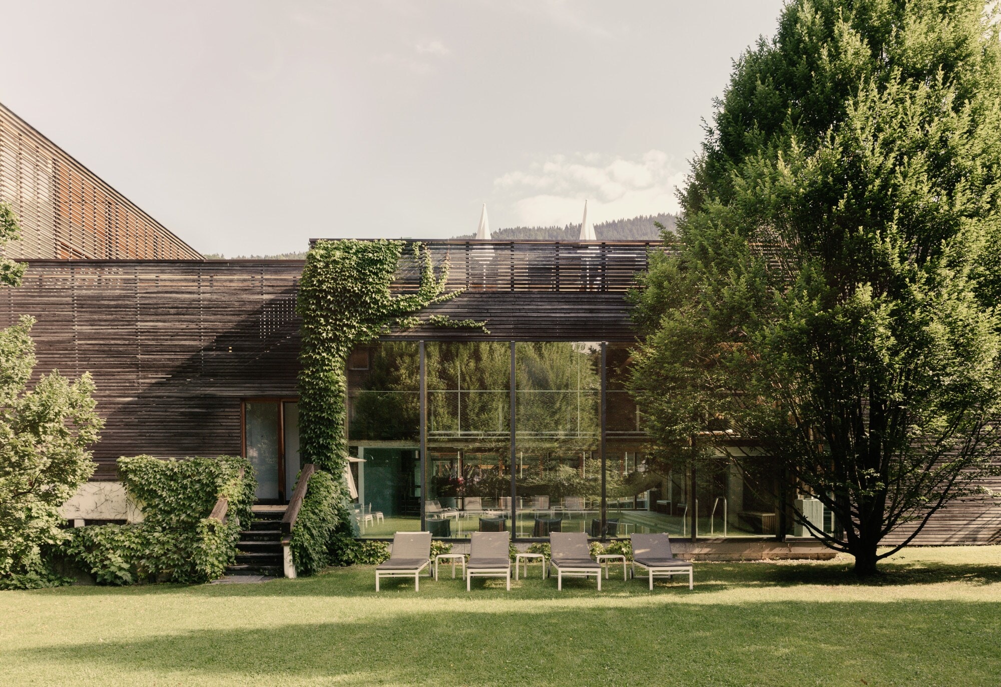 Blick auf ein modernes, holzverkleidetes Gebäude in dessen Inneren ein Swimmingpool zu sehen ist, im Vordergrund Liegestühle und Grünfläche. Blick auf ein modernes, holzverkleidetes Gebäude in dessen Inneren ein Swimmingpool zu sehen ist, im Vordergrund Liegestühle und Grünfläche.