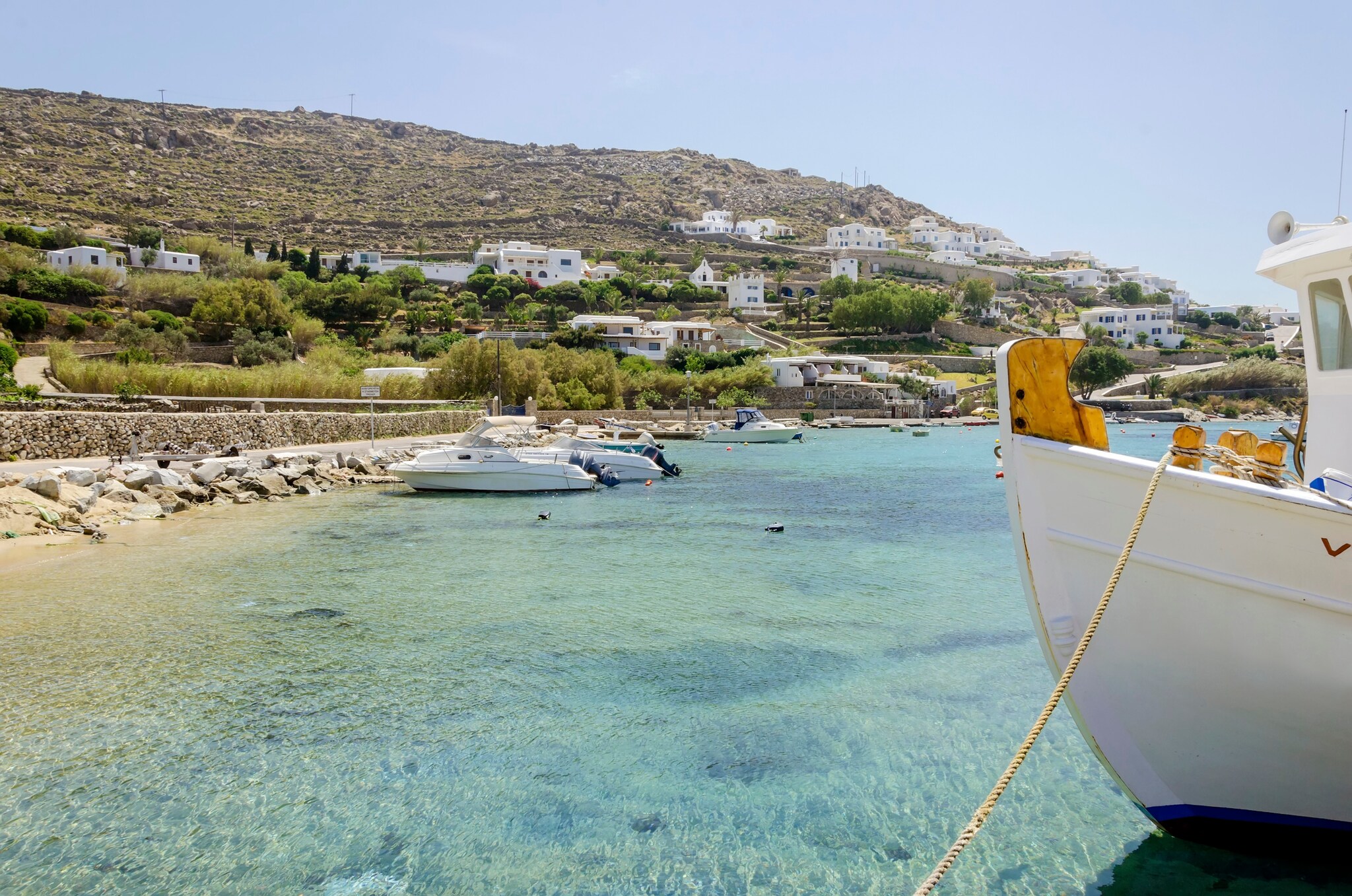 Fischerboote liegen an einem Strand von Mykonos, im Hintergrund weiß getünchte Häuser am Hügelhang