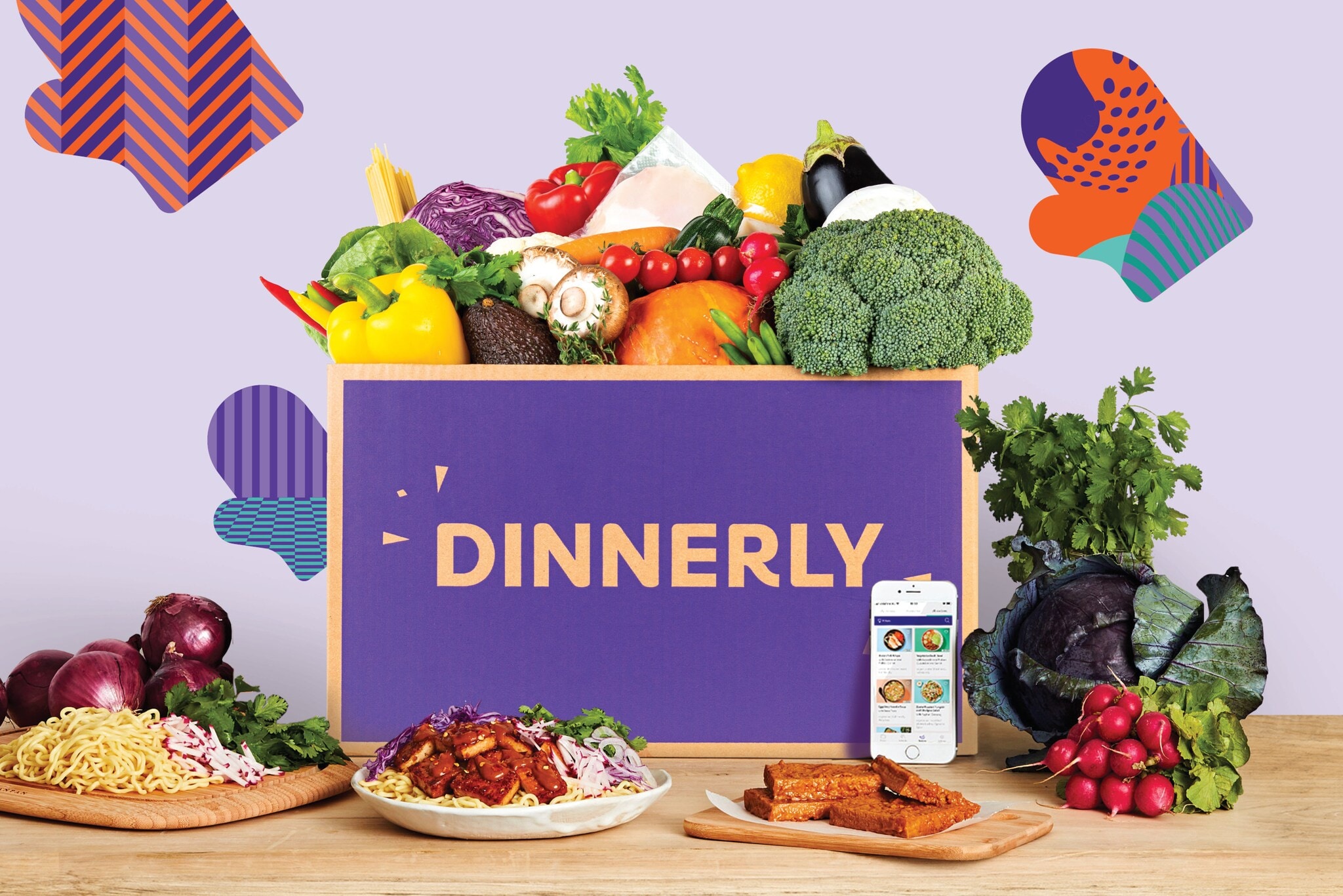 In der Mitte ein Schild mit dem Logo von Dinnerly, darüber viel Gemüse, auf dem Tisch davor diverse Gerichte auf Tellern In der Mitte ein Schild mit dem Logo von Dinnerly, darüber viel Gemüse, auf dem Tisch davor diverse Gerichte auf Tellern