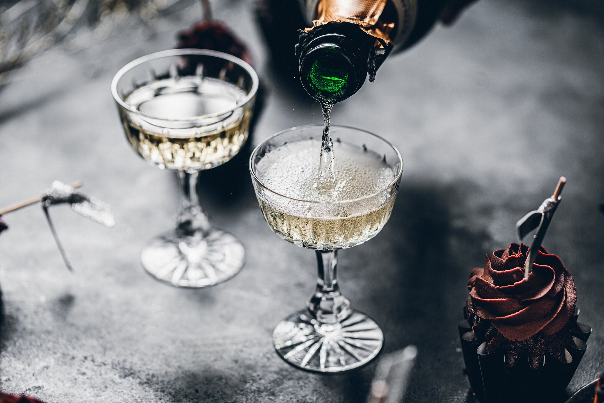 Zwei Gläser auf einem grauen Tisch werden mit Champagner gefüllt, daneben ist ein Cupcake mit Schokohaube zu sehen Zwei Gläser auf einem grauen Tisch werden mit Champagner gefüllt, daneben ist ein Cupcake mit Schokohaube zu sehen