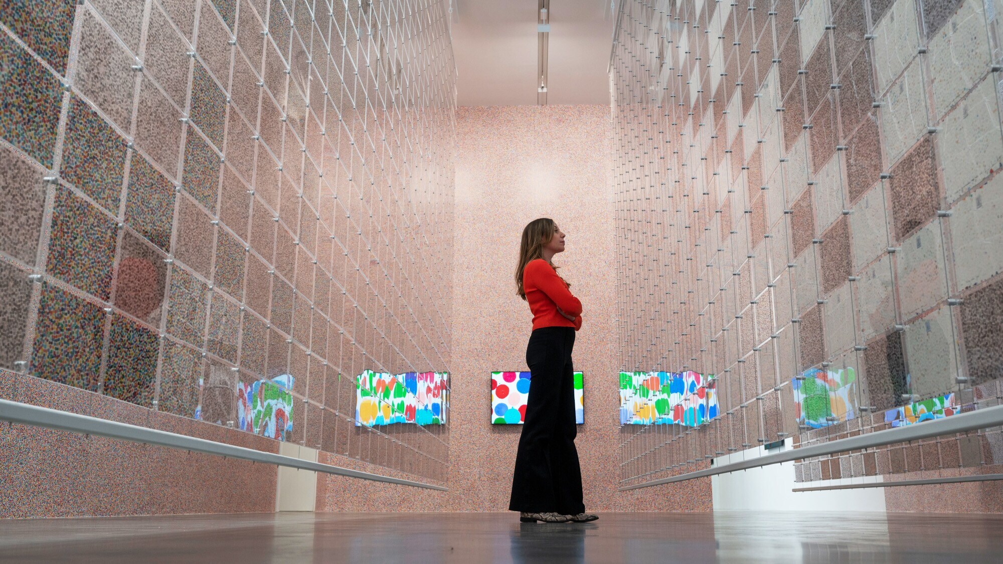 Eine Frau betrachtet gepunktete Kunstwerke hinter einer hängenden Plexiglaswand in einer Galerie, im Hintergrund digitale Kunstwerke auf Monitoren