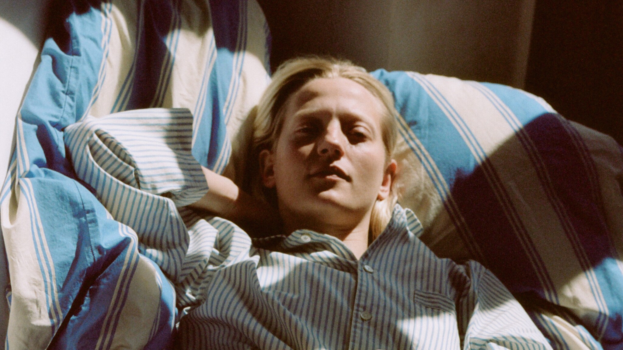 Porträt einer jungen Frau, die in einem gestreiften Pyjama auf gestreifter Bettwäsche im Sonnenlicht liegt