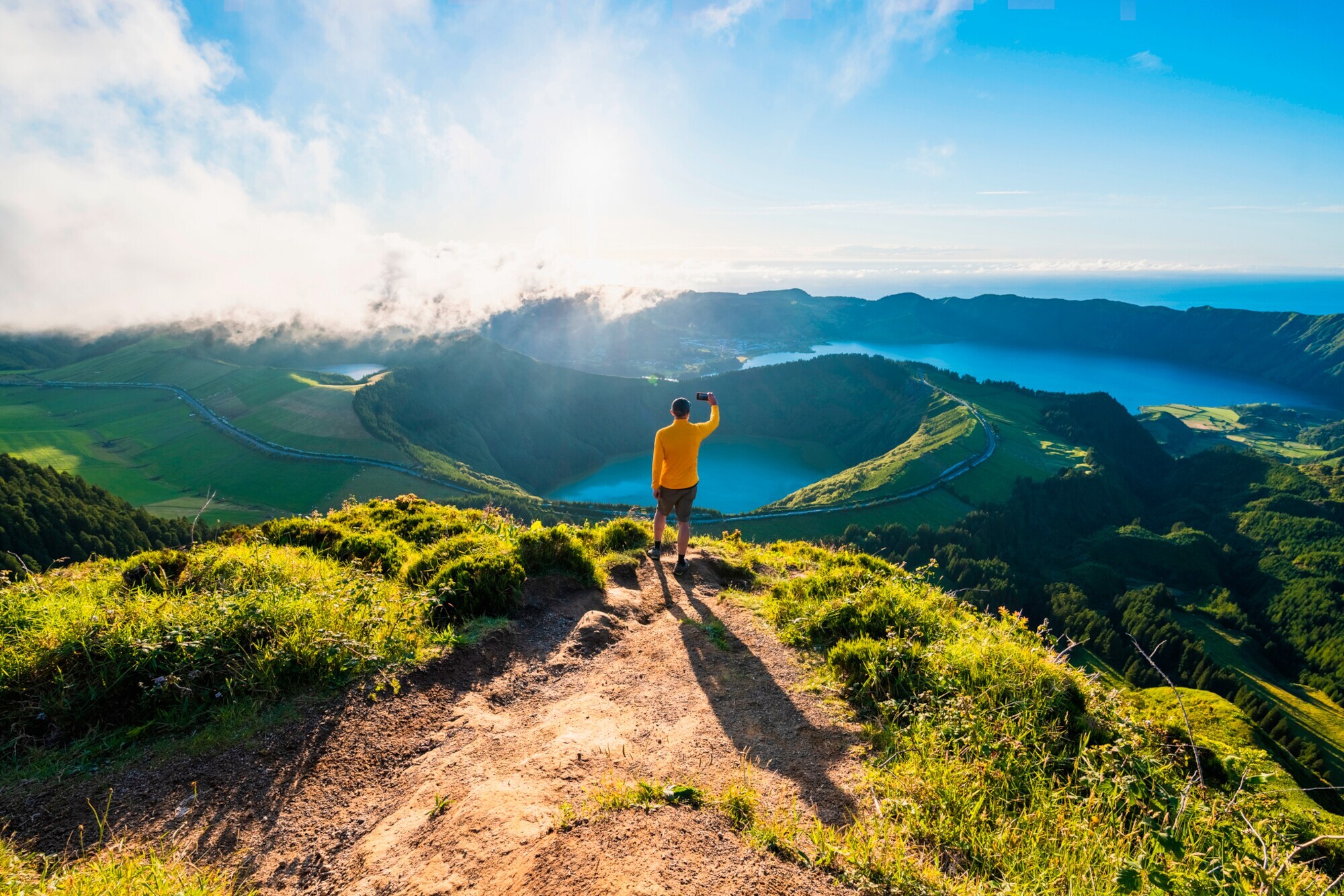 Eine Person im gelben Oberteil steht auf einem Berg und blickt auf einen Vulkan auf den Azoren