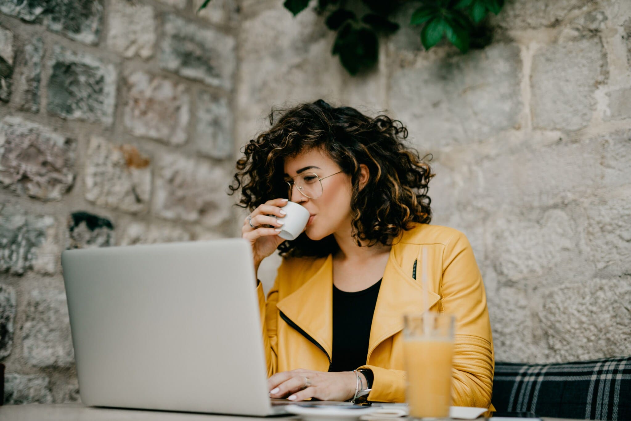 Eine Frau in gelber Jacke mit lockigen schwarzen Haaren und Brille sitzt im Café am Laptop und trinkt einen Espresso Eine Frau in gelber Jacke mit lockigen schwarzen Haaren und Brille sitzt im Café am Laptop und trinkt einen Espresso