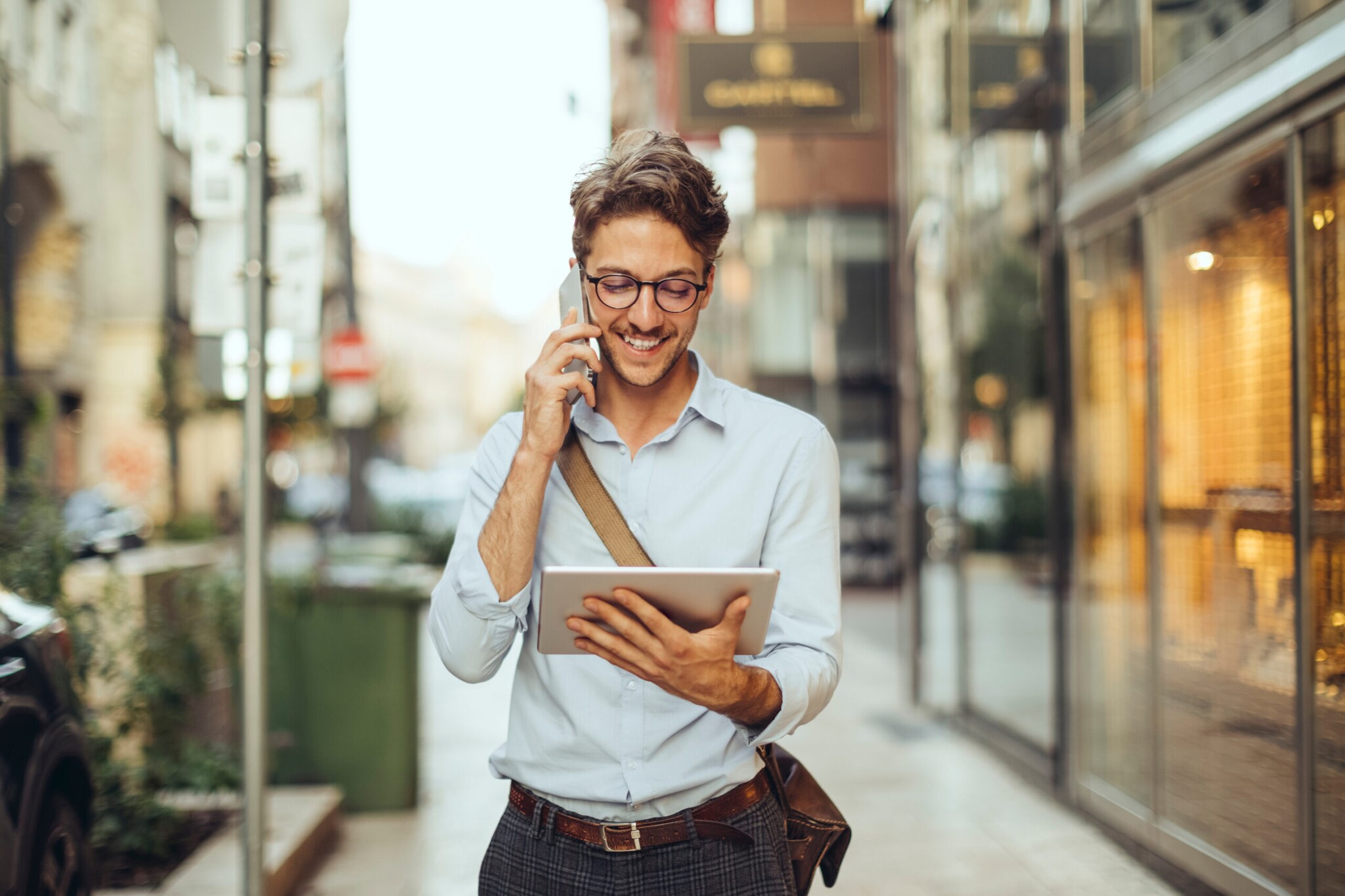 Ein junger Mann mit Brille im weißen Hemd telefoniert und schaut dabei auf ein Tablet