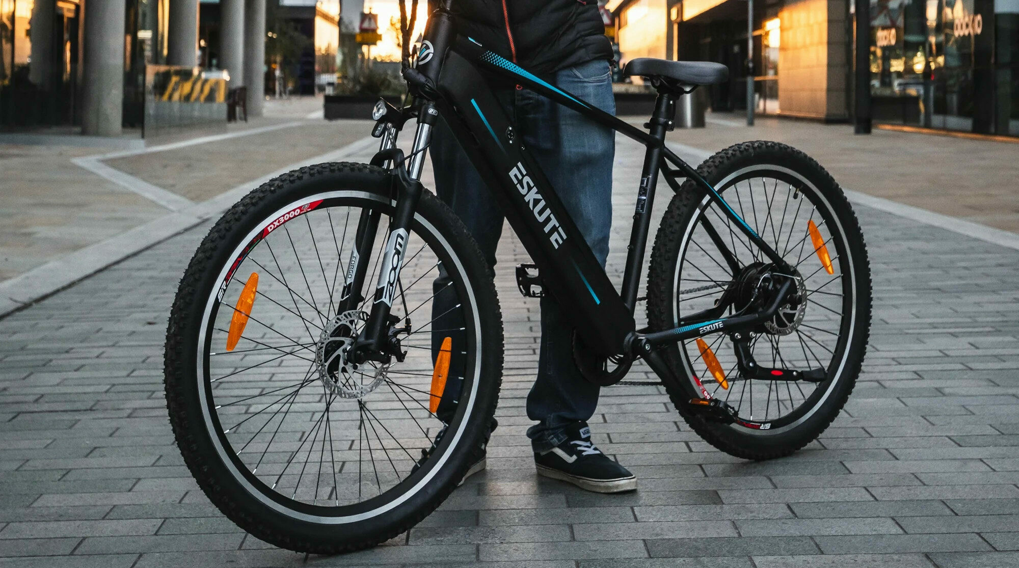 Ein Mann steht hinter einem dunklen E-Bike, rechts und links sind hohe Gebäude zu sehen Ein Mann steht hinter einem dunklen E-Bike, rechts und links sind hohe Gebäude zu sehen