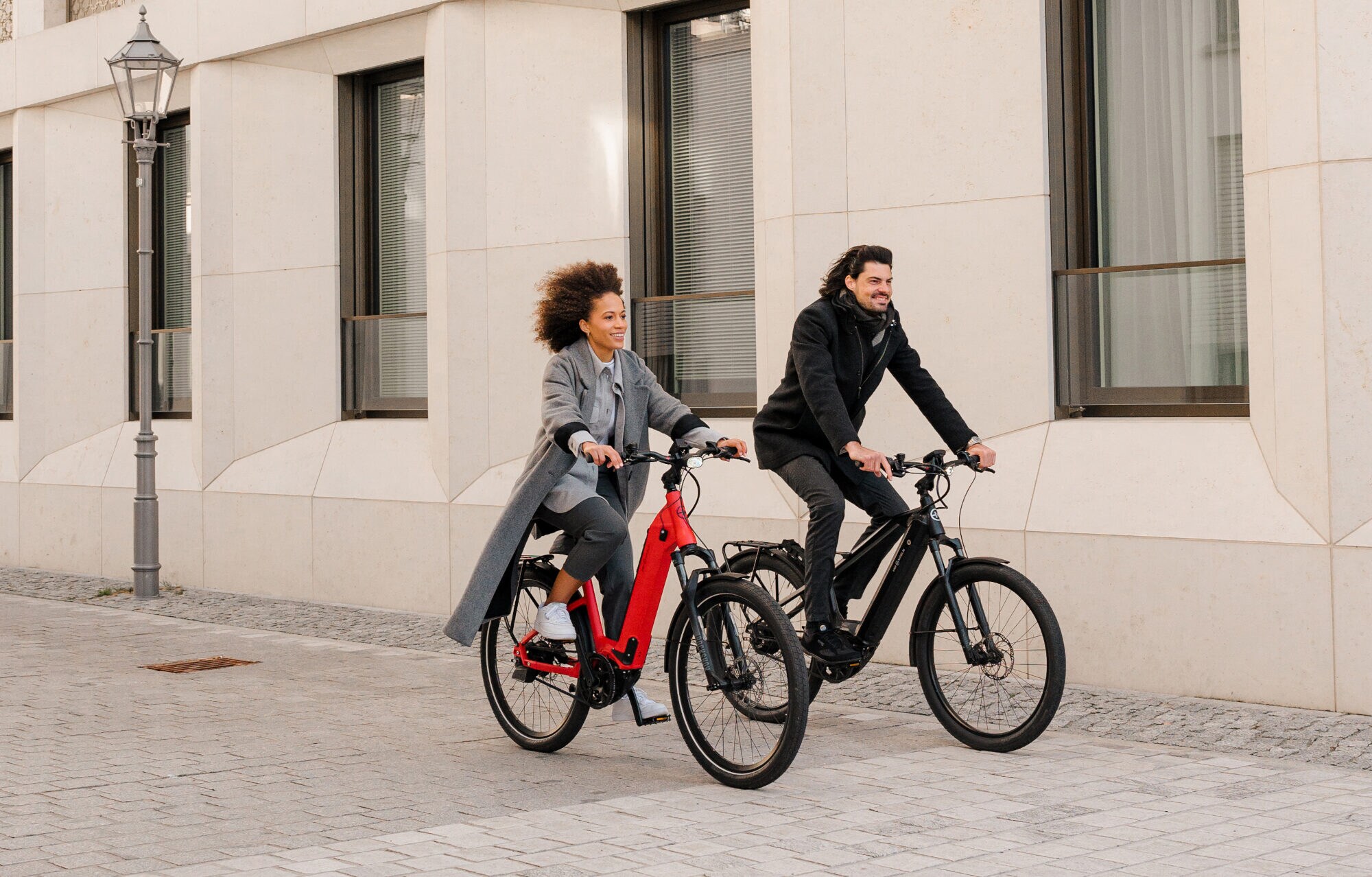 Ein Mann und eine Frau radeln auf zwei E-Bikes an einem Haus vorbei Ein Mann und eine Frau radeln auf zwei E-Bikes an einem Haus vorbei