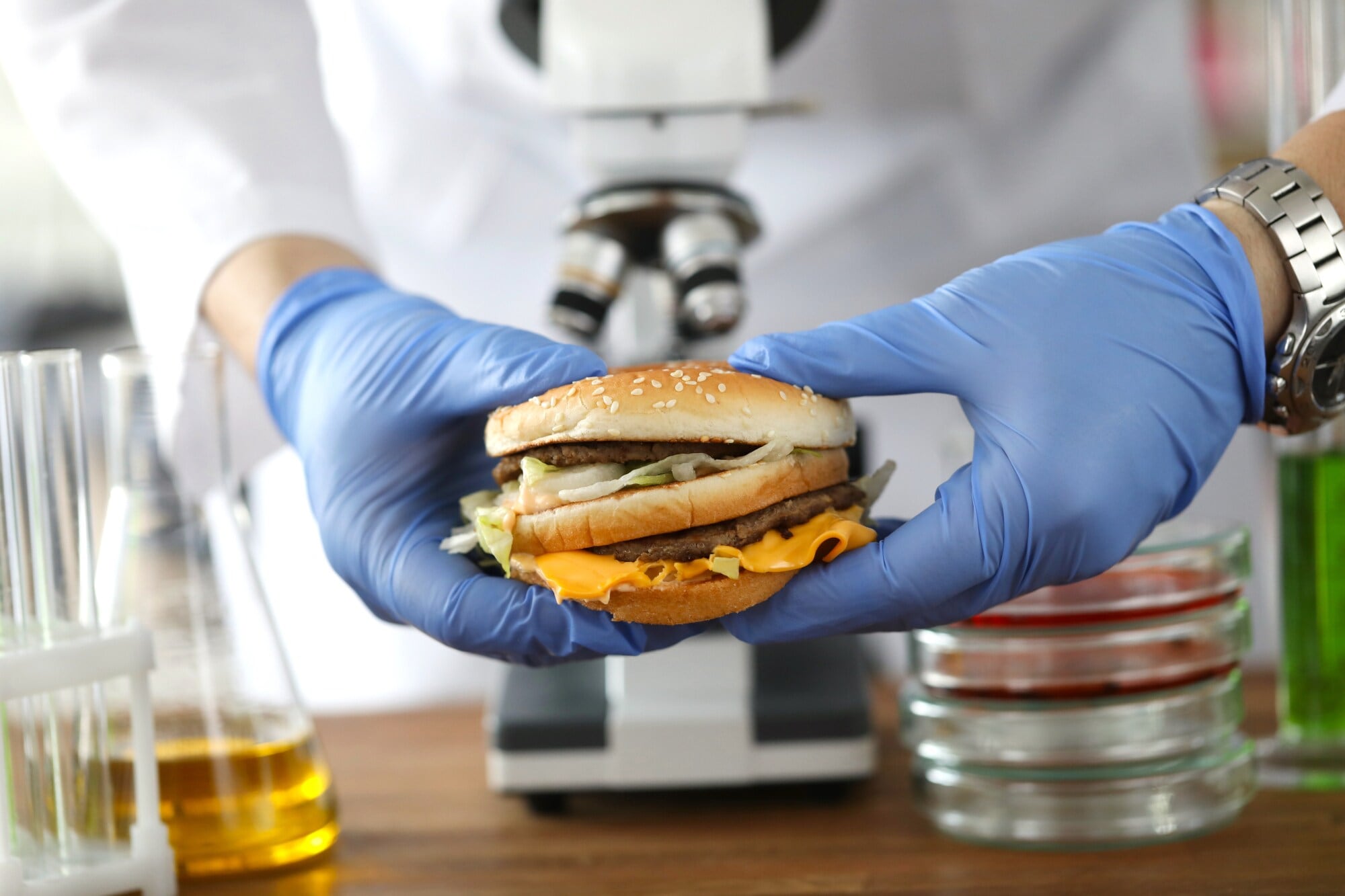 Eine Person im weißen Kittel mit blauen Gummihandschuhen hält einen Burger vor einem Mikroskop fest Eine Person im weißen Kittel mit blauen Gummihandschuhen hält einen Burger vor einem Mikroskop fest