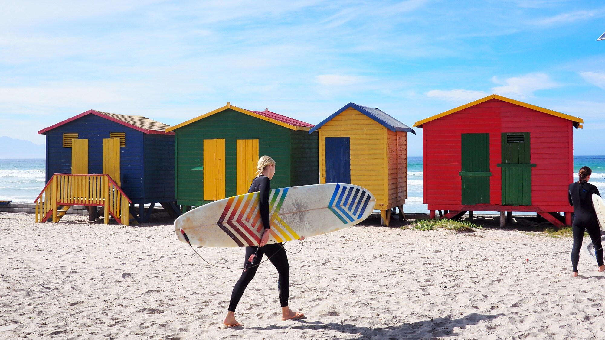 Surferinnen vor bunten Umkleidekabinen aus Holz an einem Strand bei Kapstadt