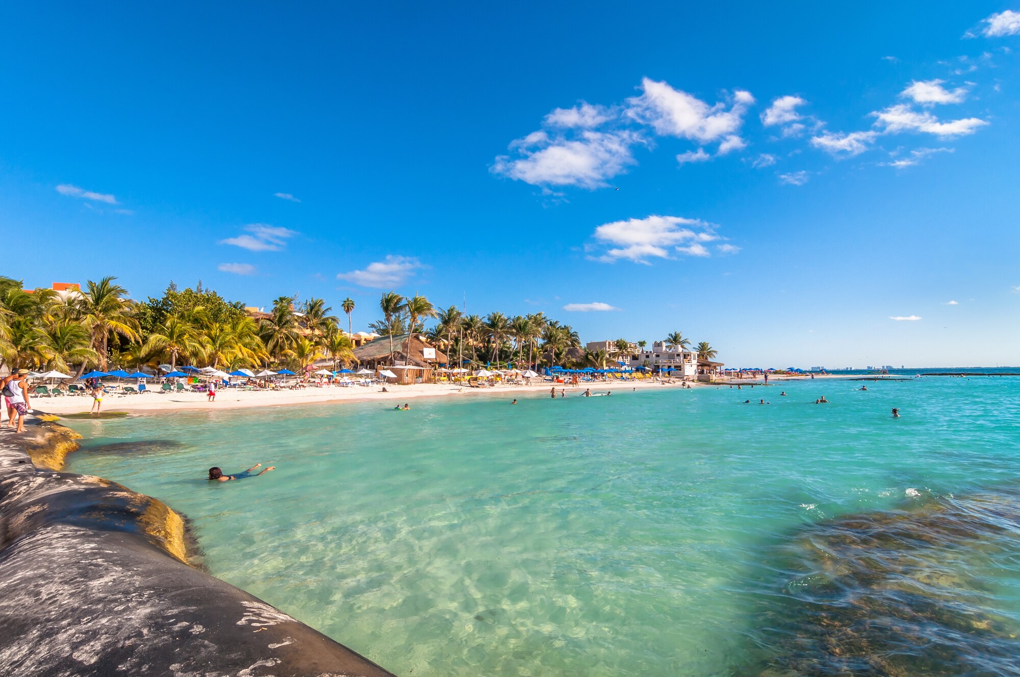 Badende im flachen Wasser an einem tropischen, touristischen Sandstrand mit Palmen und Badeliegen