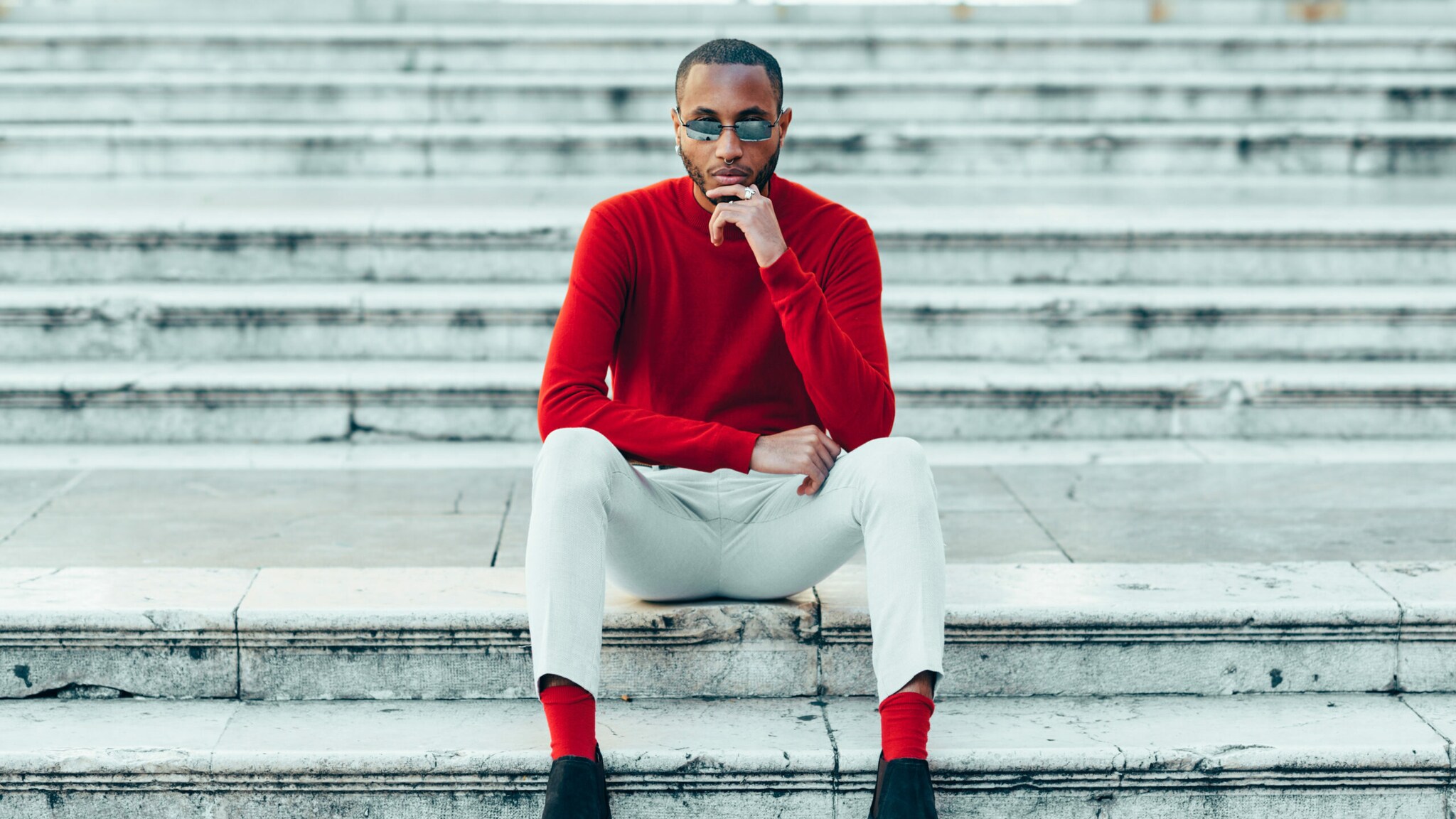 Ein Mann in weißen Hosen und einem roten Kaschmirpullover sitzt auf einer weißen Marmirtreppe