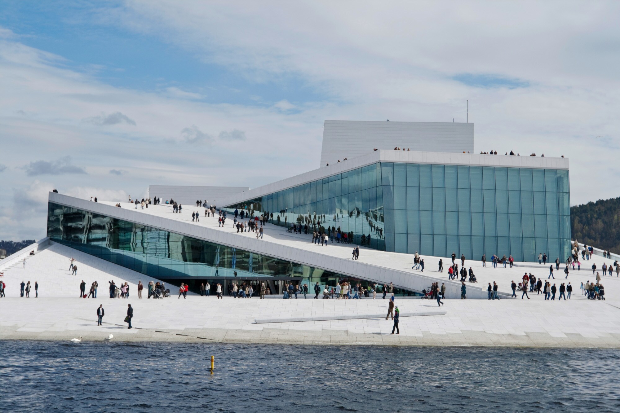 Personen laufen auf einem weißen, futuristischen Gebäude mit Glasfassade am Wasser