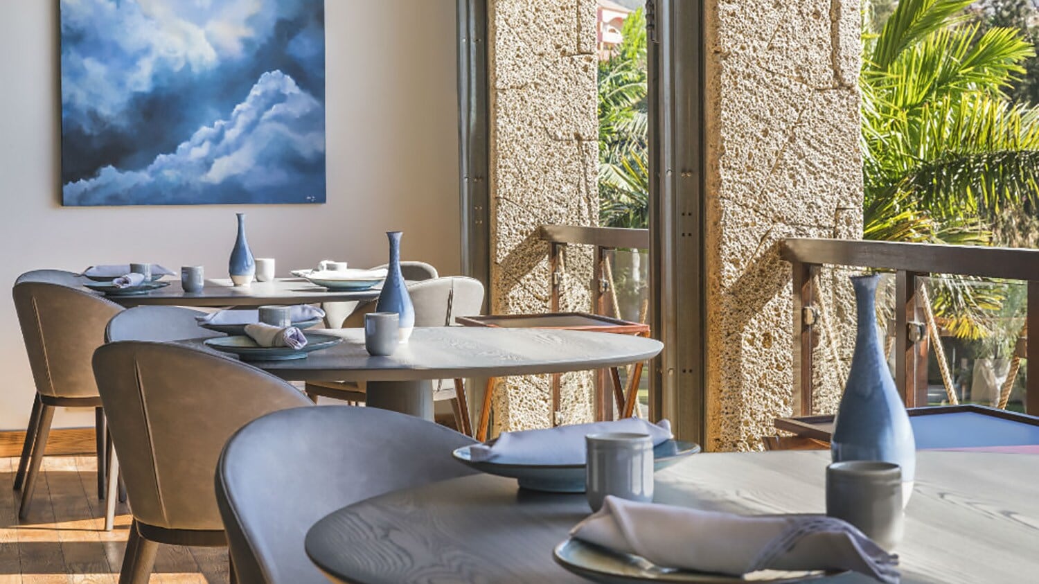 Innenraum eines luftigen, hellen Restaurants mit gedeckten Tischen an einer Fensterfront mit Blick auf Palmen Innenraum eines luftigen, hellen Restaurants mit gedeckten Tischen an einer Fensterfront mit Blick auf Palmen