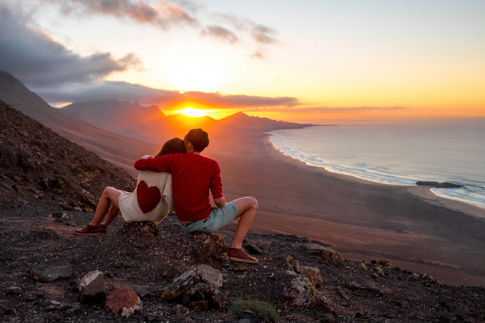 Zwei Personen sitzen mit dem Rücken zur Kamera auf einem Felshang und Blicken in den Sonnenuntergang über den Bergen, rechts im Bild das Meer