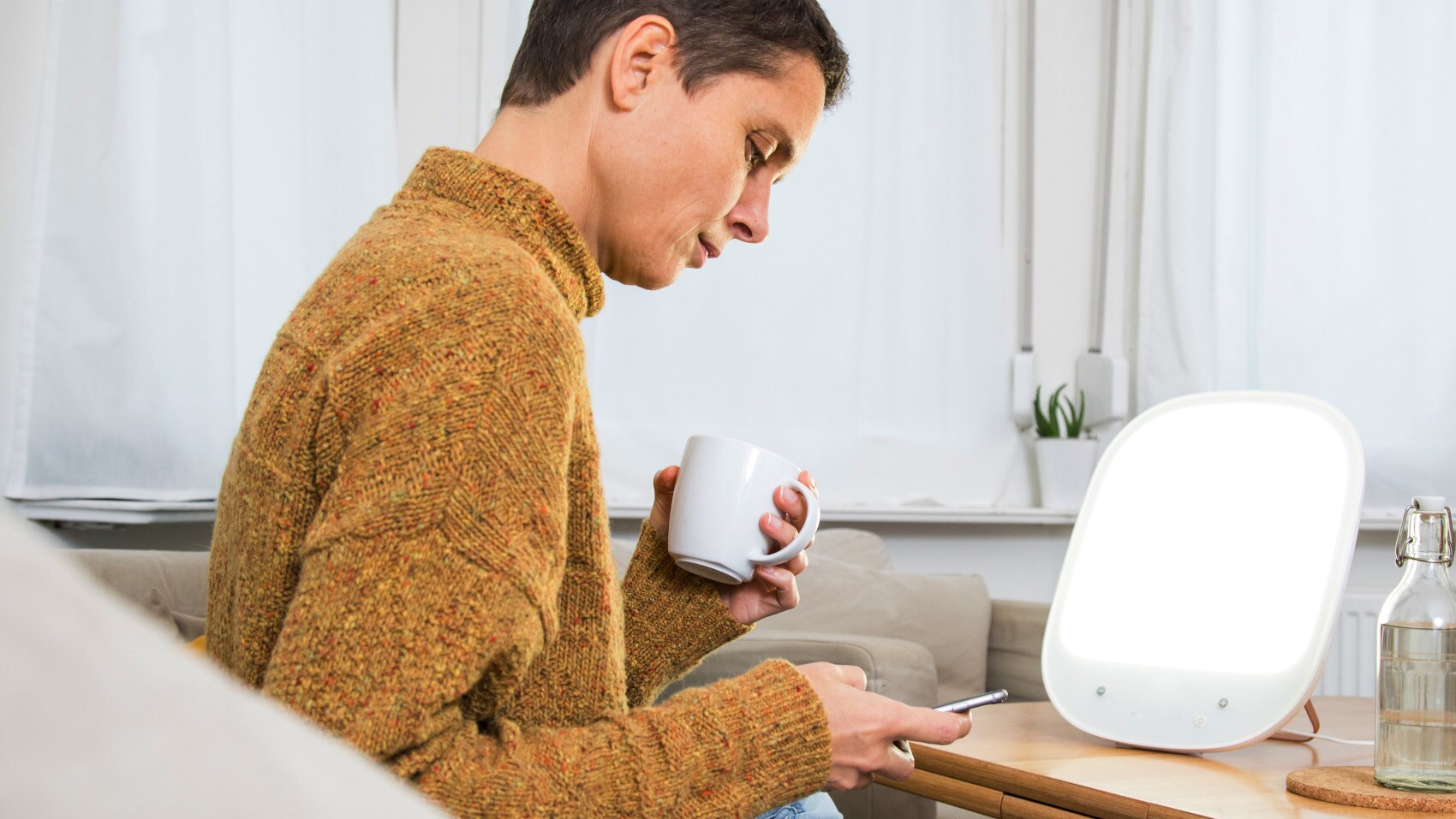 Eine Frau sitzt im Wohnzimmer vor einer Tageslichtlampe und bedient ihr Smartphone