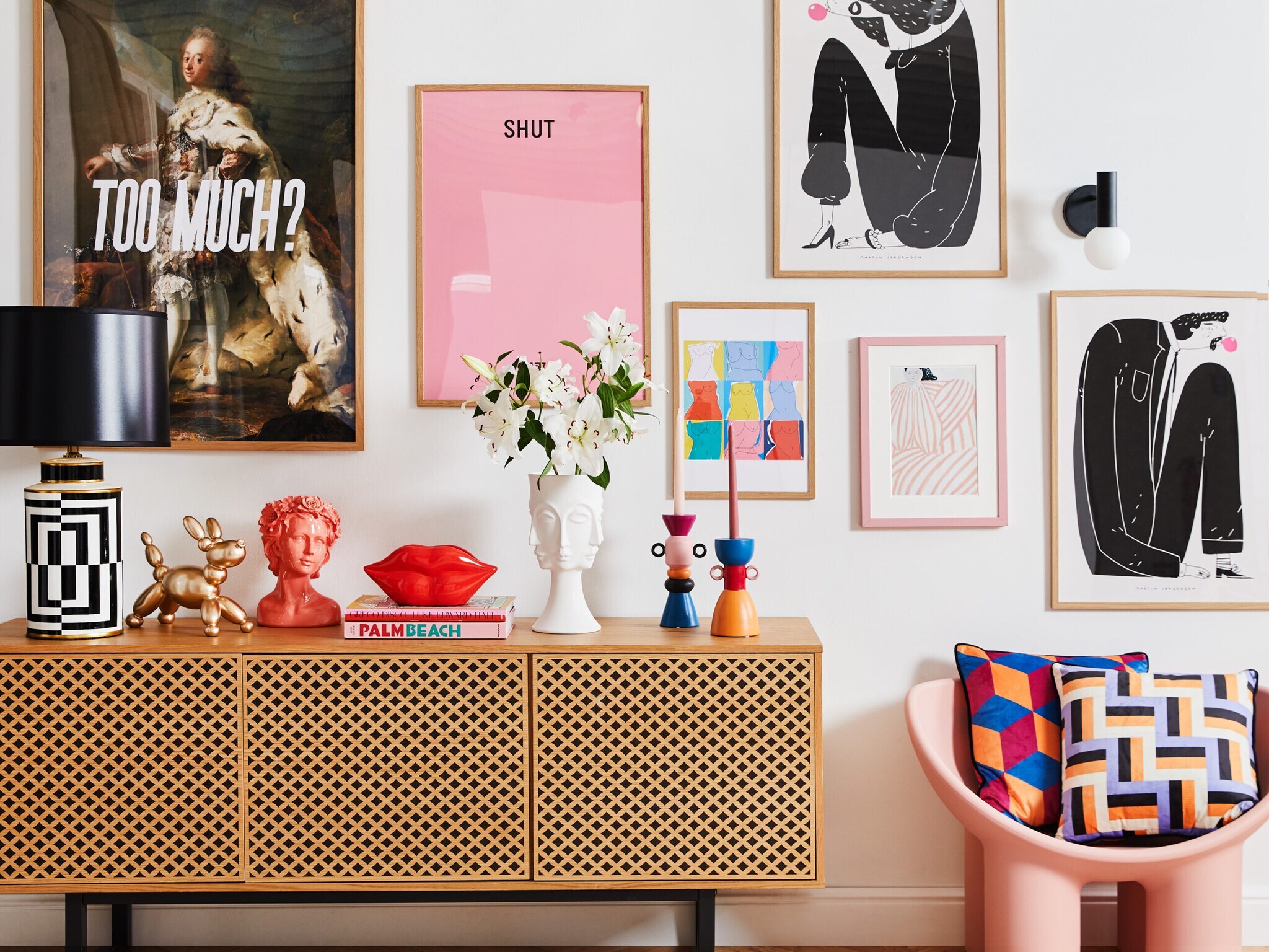 Eine gemusterte Holzkommode mit verspielten Accessoires obendrauf und ein rosa Stuhl mit gemusterten Kissen vor einer Wand mit gerahmten Bildern