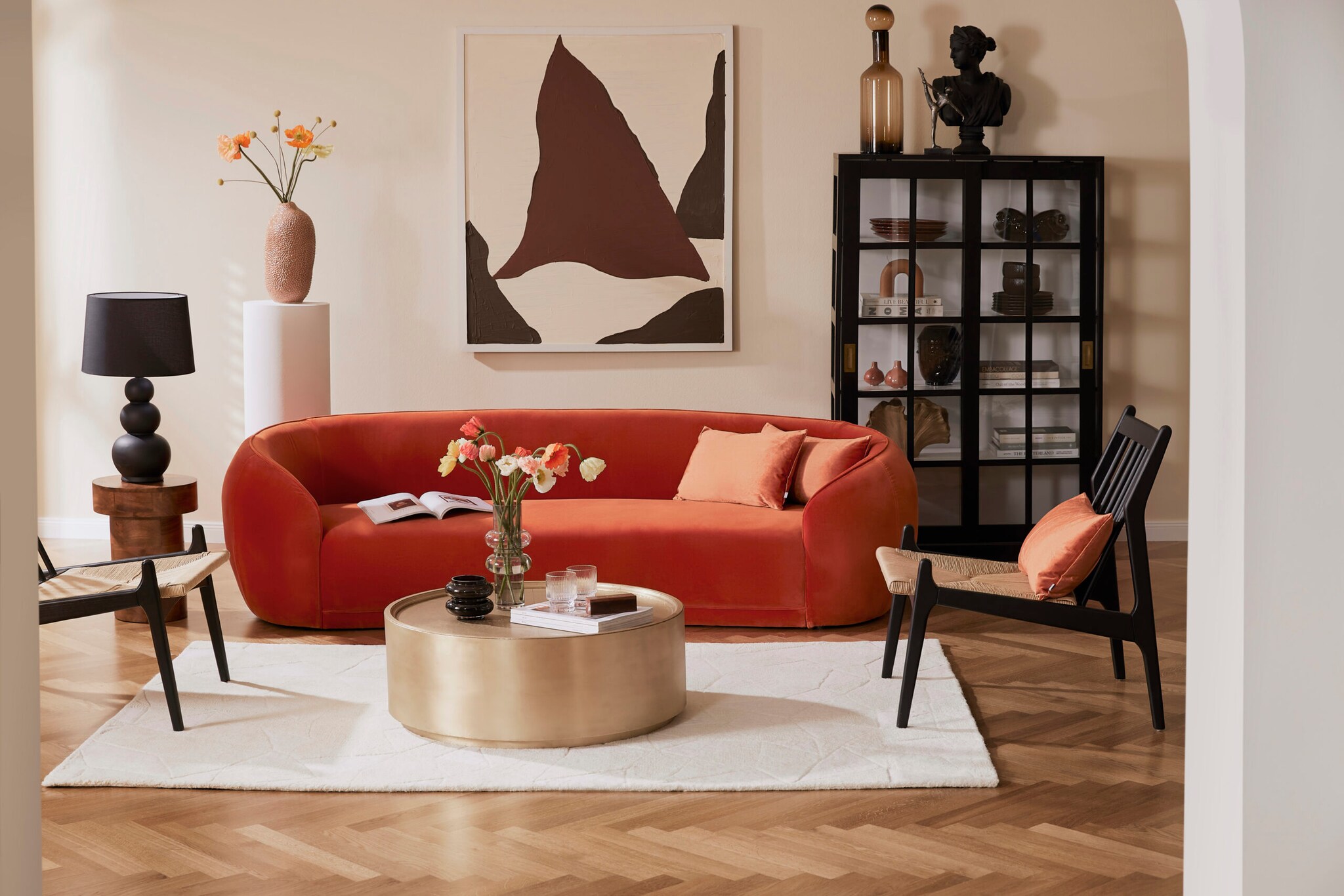 Ein modernes, helles Wohnzimmer mit einem geschwungenen, orangefarbenen Samtsofa vor einem runden, goldenen Couchtisch, daneben schwarze Möbelstücke als Akzent Ein modernes, helles Wohnzimmer mit einem geschwungenen, orangefarbenen Samtsofa vor einem runden, goldenen Couchtisch, daneben schwarze Möbelstücke als Akzent