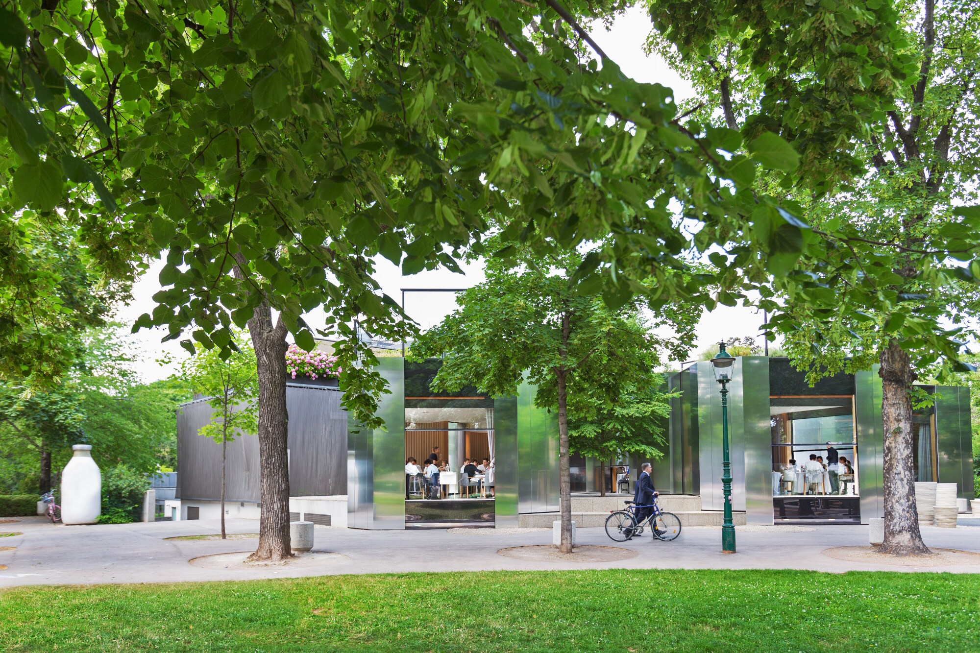 Rendering des Wiener Stadtparks mit Bäumen und Wegen, im Hintergrund ein Restaurant mit offenen Türen und Gästen im Inneren Rendering des Wiener Stadtparks mit Bäumen und Wegen, im Hintergrund ein Restaurant mit offenen Türen und Gästen im Inneren