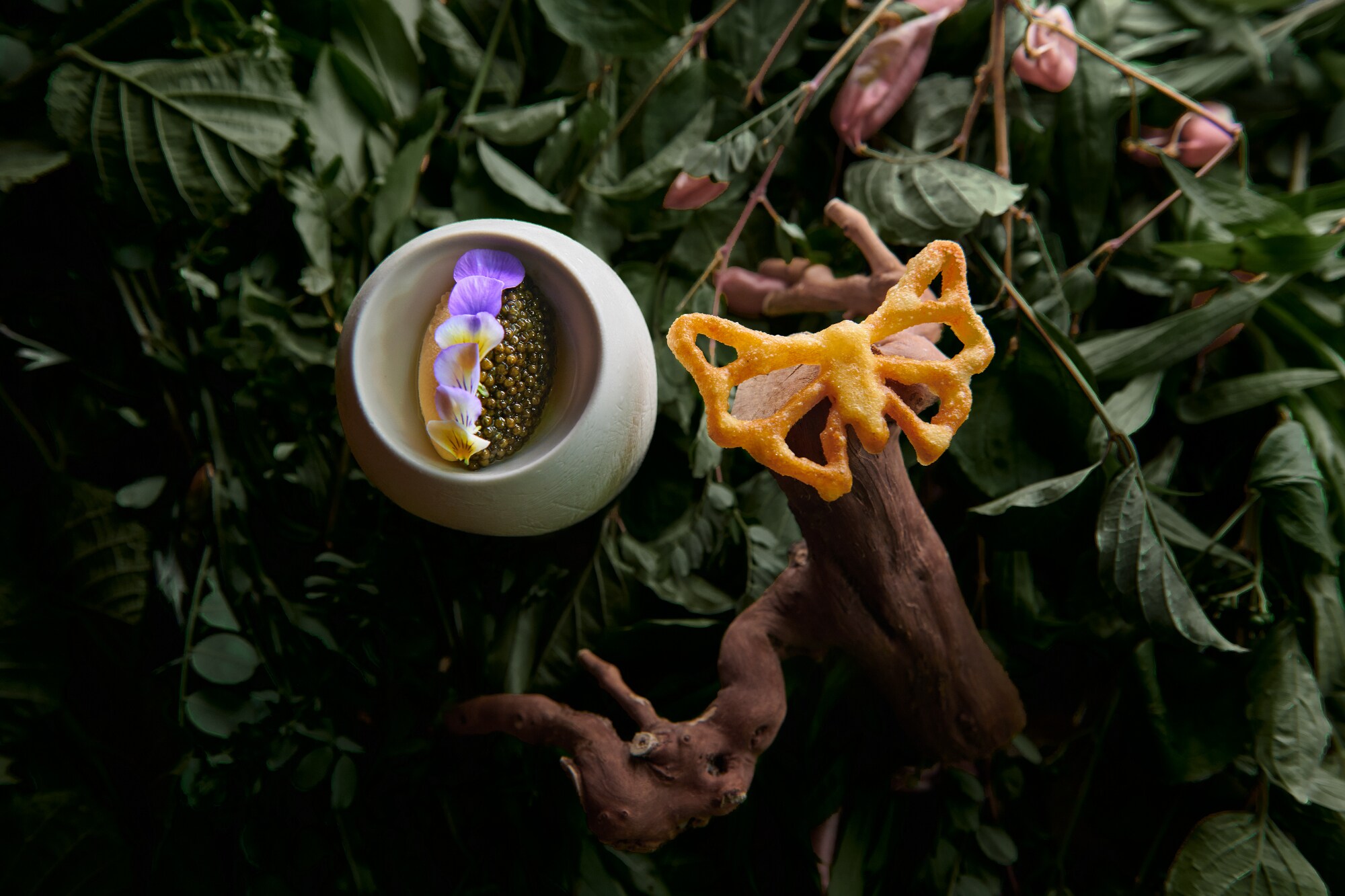 Ein aus Teig geformter frittierter Schmetterling und eine Schüssel mit Essen und Blumen sind auf Blättern und Blumen arrangiert