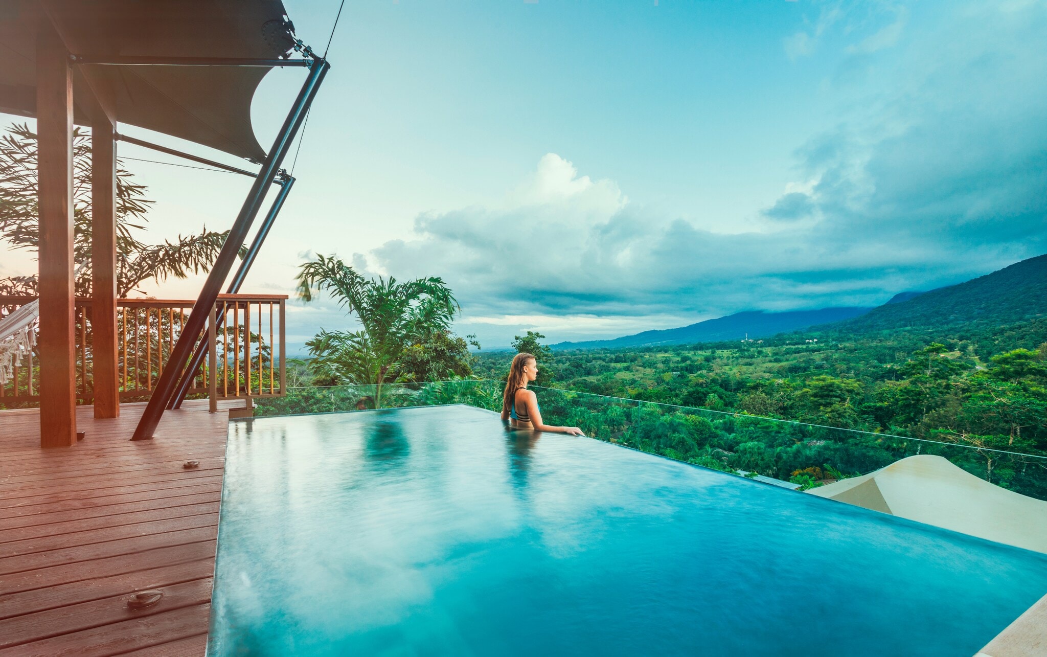 Profilansicht einer Frau, die aus einem Infinity-Pool über eine tropische Hügellandschaft schaut Profilansicht einer Frau, die aus einem Infinity-Pool über eine tropische Hügellandschaft schaut