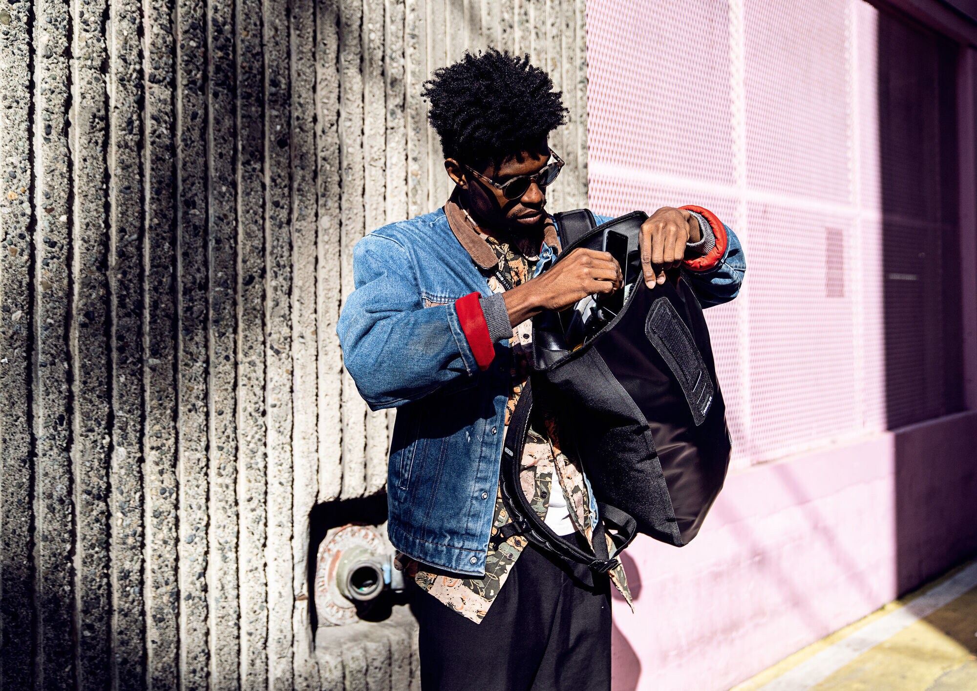 Ein Mann mit Sonnenbrille packt sein Smartphone in einen über einer Schulter getragenen Laptop-Rucksack Ein Mann mit Sonnenbrille packt sein Smartphone in einen über einer Schulter getragenen Laptop-Rucksack