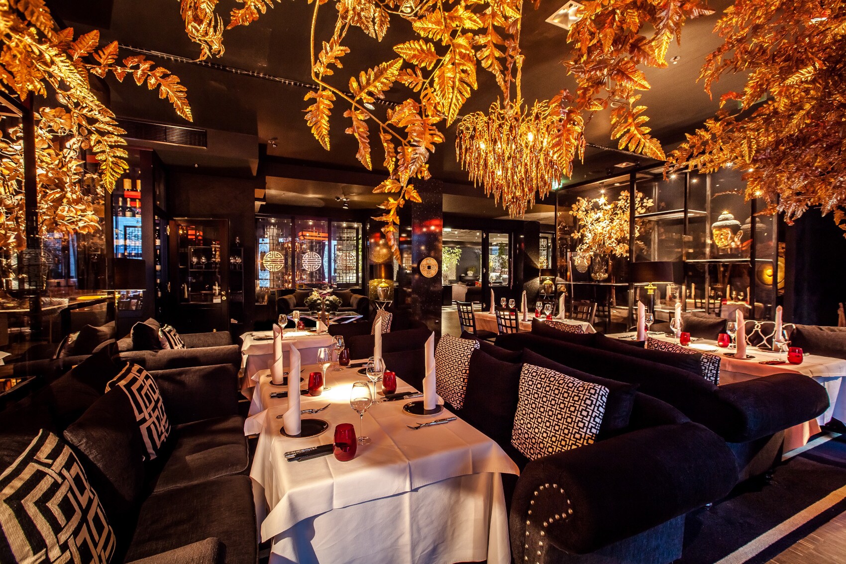 Ein Restaurant mit mehreren, weiß gedeckten Tischen, dunklen Sitzmöbeln und goldenen Leuchtern und Dekoblättern. Ein Restaurant mit mehreren, weiß gedeckten Tischen, dunklen Sitzmöbeln und goldenen Leuchtern und Dekoblättern.