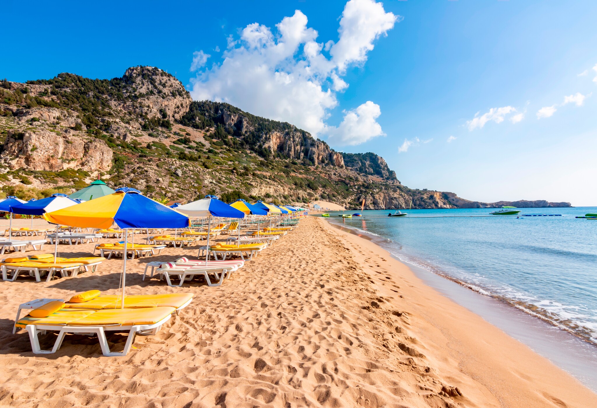 Liegen und gelbblaue Sonnenschirme an einem Strand, im Hintergrund Berge Liegen und gelbblaue Sonnenschirme an einem Strand, im Hintergrund Berge