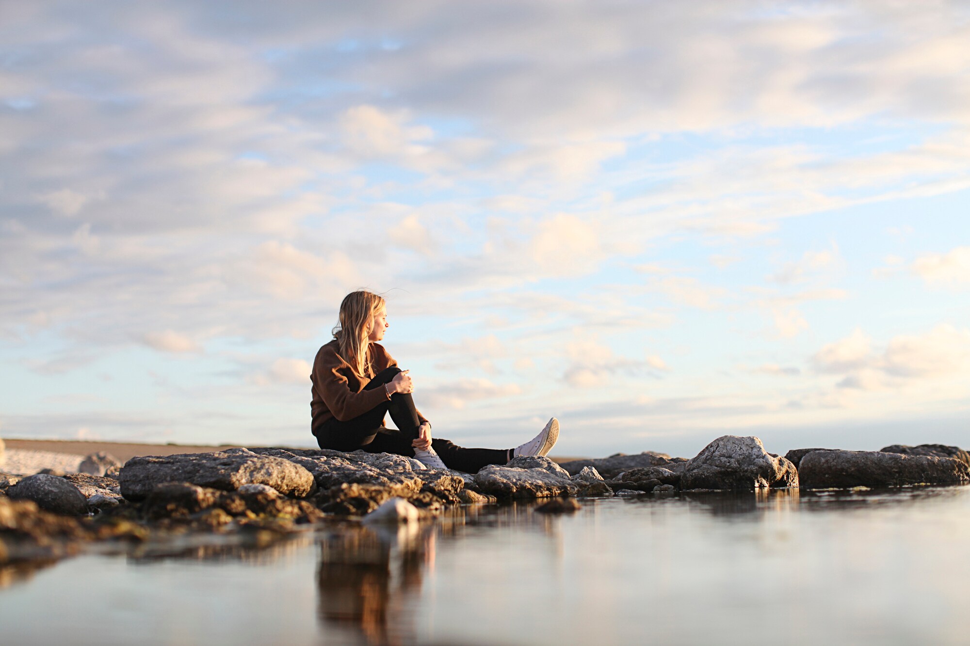 Junge Frau mit blonden langen Haaren sitzt auf Felsen und schaut übers Meer