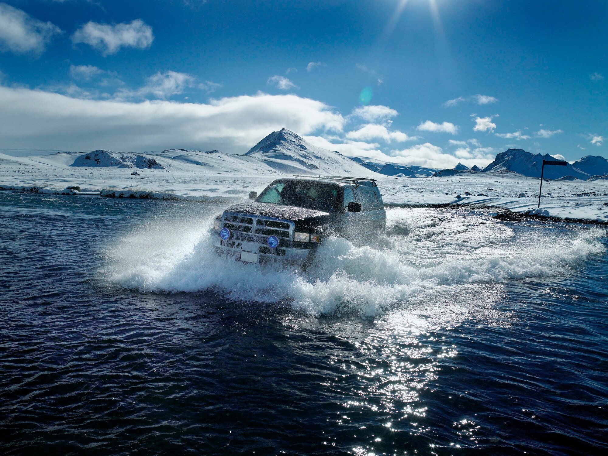 Ein Fahrzeug fährt durch einen Fluss, dahinter erstreckt sich eine Schneelandschaft.