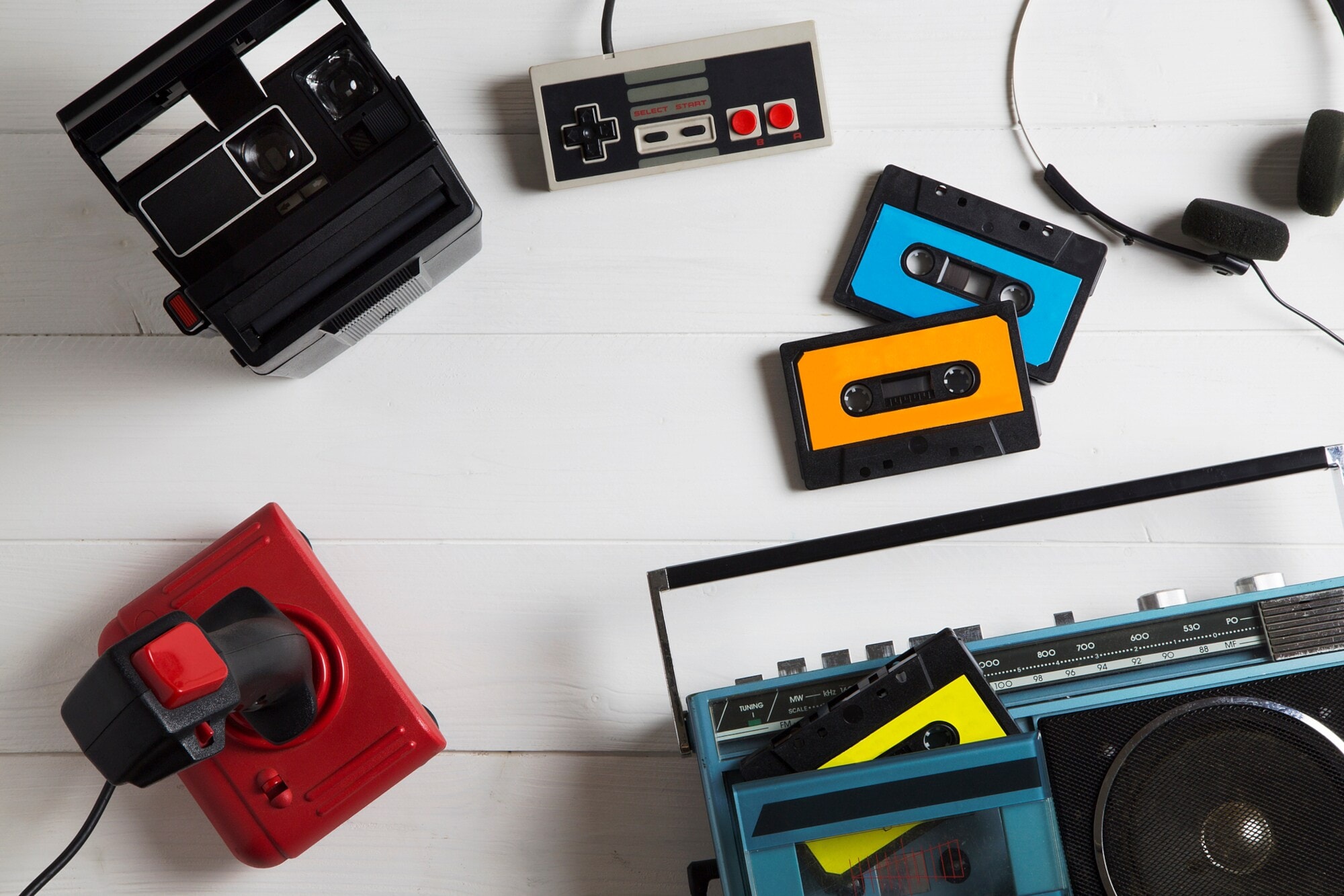 Auf einem Tisch liegen mehrere analoge Medien in Form von Kassetten, einer Polaroidkamera, einem Kassettenrekorder, einem NES-Gamepad und Joystick Auf einem Tisch liegen mehrere analoge Medien in Form von Kassetten, einer Polaroidkamera, einem Kassettenrekorder, einem NES-Gamepad und Joystick