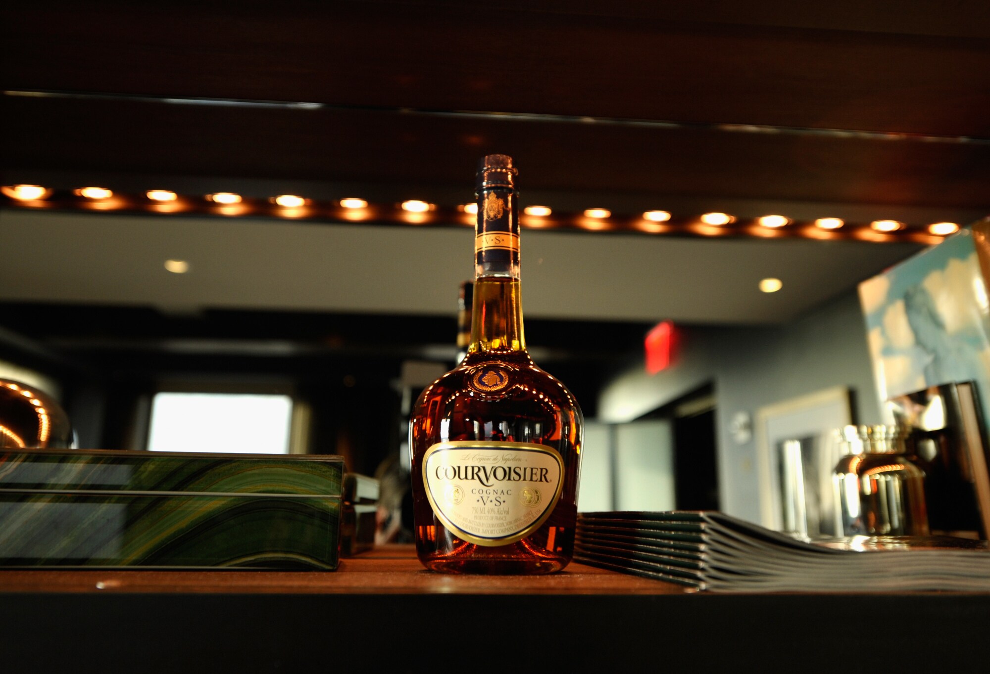 Eine Flasche Courvoisier steht neben einem Heftstapel und einer Box auf einem Tisch. Im Hintergrund hängt ein beleuchteter Spiegel