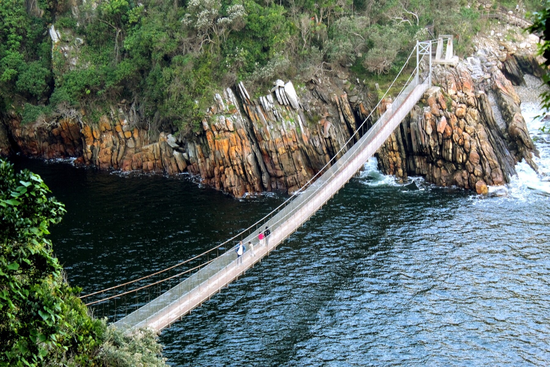 Personen auf einer Hängebrücke zwischen bewaldeten Felsklippen über einer Meeresmündung