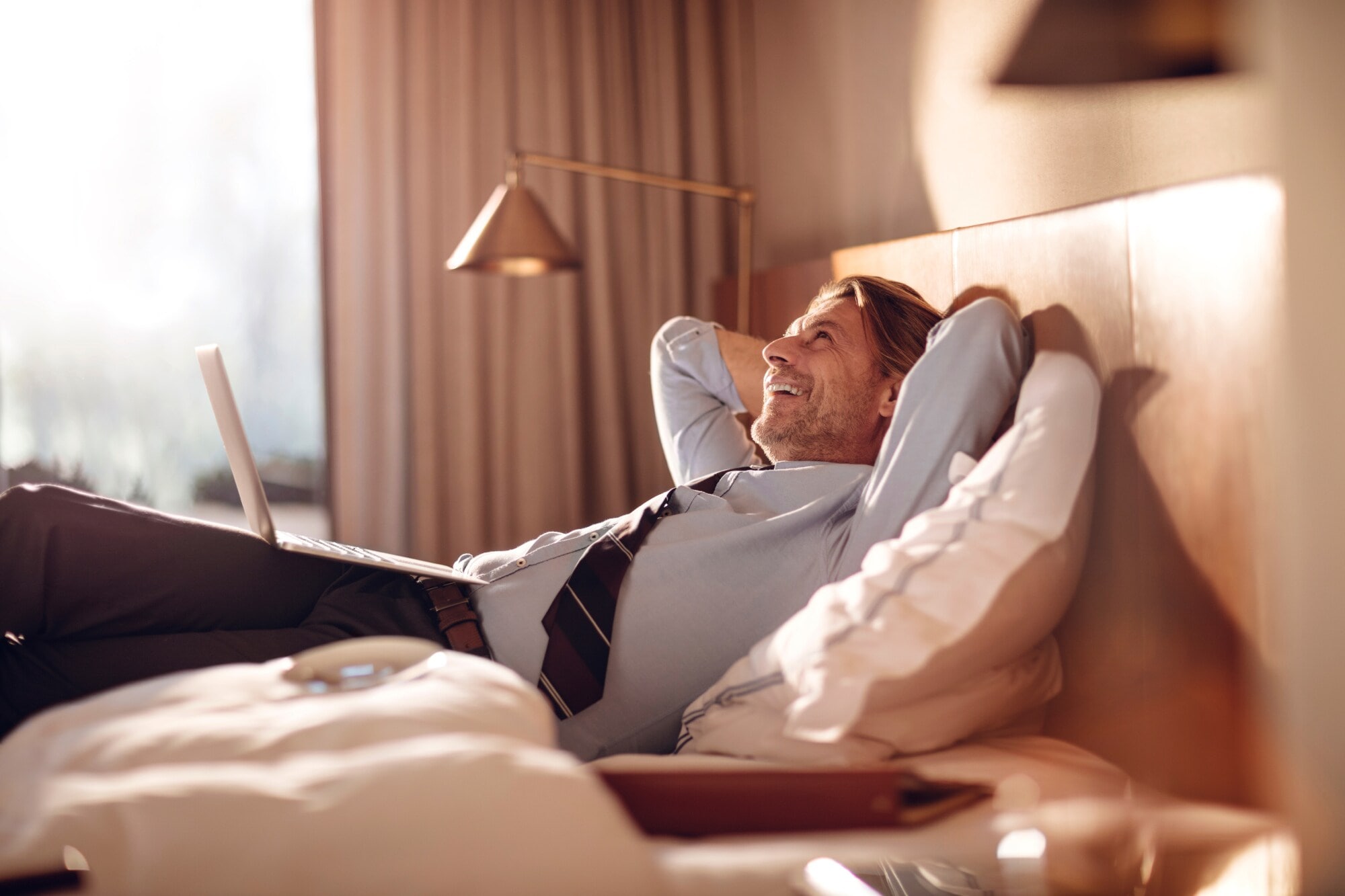 Ein Geschäftsmann liegt entspannt mit einem Laptop in einem Hotelbett Ein Mann mit Hose, Hemd und Krawatte liegt lächelnd und mit hinter dem Kopf verschränkten Armen mit einem Laptop in einem Hotelbett.