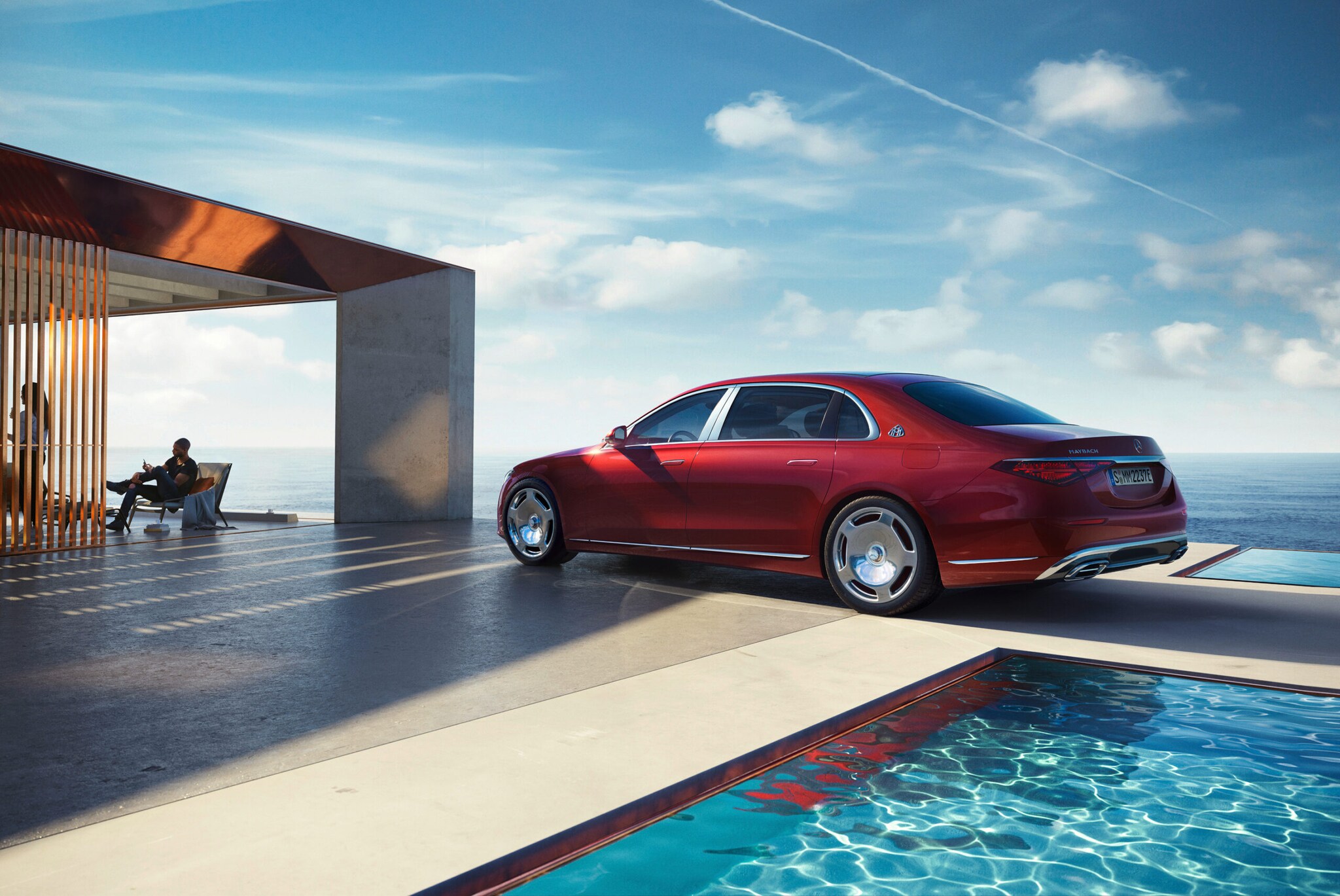 Ein roter Mercedes-Maybach von der Seite am Pool eines modernen Hauses, im Hintergrund Meer und Personen im Haus