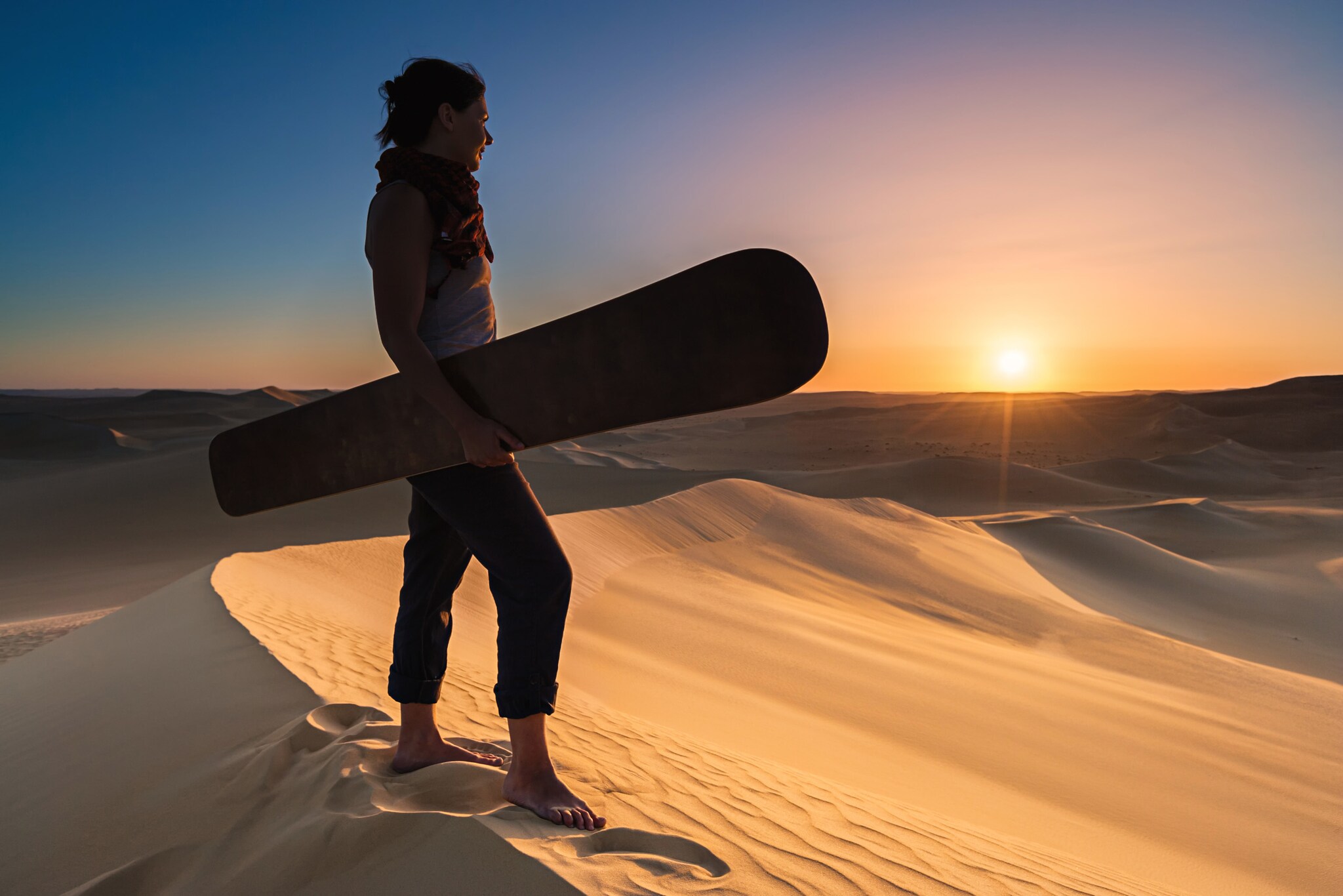 Eine Frau mit einem Sandboard unter dem Arm blickt von einer Sanddüne über eine Wüstenlandschaft in den Sonnenuntergang
