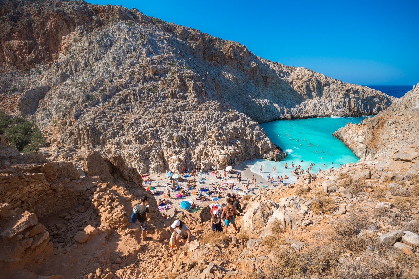 Blick auf einen zwischen Felsen liegenden Sandstrand, der einen schmalen Zugang zum türkisfarbenen Meer bietet