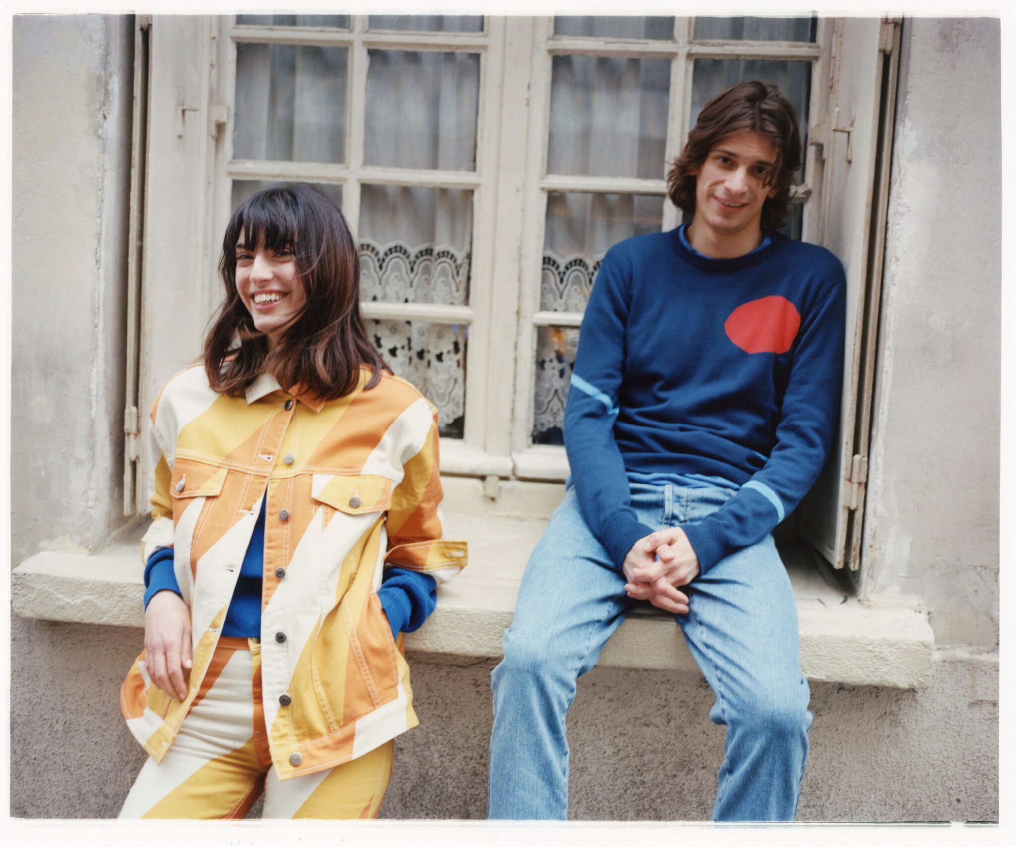Ein Mann und eine Frau vor einem Fenster eines alten Gebäudes tragen farbige und lässige Outfits.